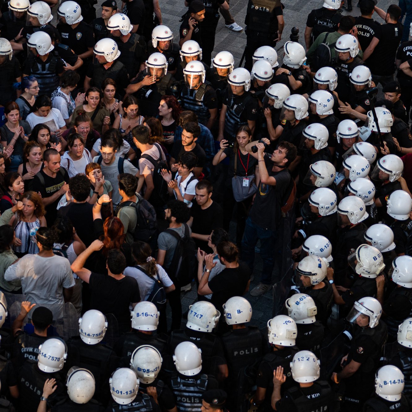 در این تصویر، گروهی از مردم در حال تظاهرات یا تجمعی هستند که با نیروهای پلیس محاصره شده‌اند. افراد معترض در وسط تصویر قرار دارند و به نظر می‌رسد در حال گفت‌وگو یا گفتگو با پلیس هستند. نیروهای پلیس در اطراف آنها ایستاده‌اند و کلاه‌های مخصوصی به سر دارند. فضای تصویر نشان‌دهنده تنش و فعالیت اجتماعی است، با تجمع جمعیتی که به سمت نیروهای پلیس نگاه می‌کنند.