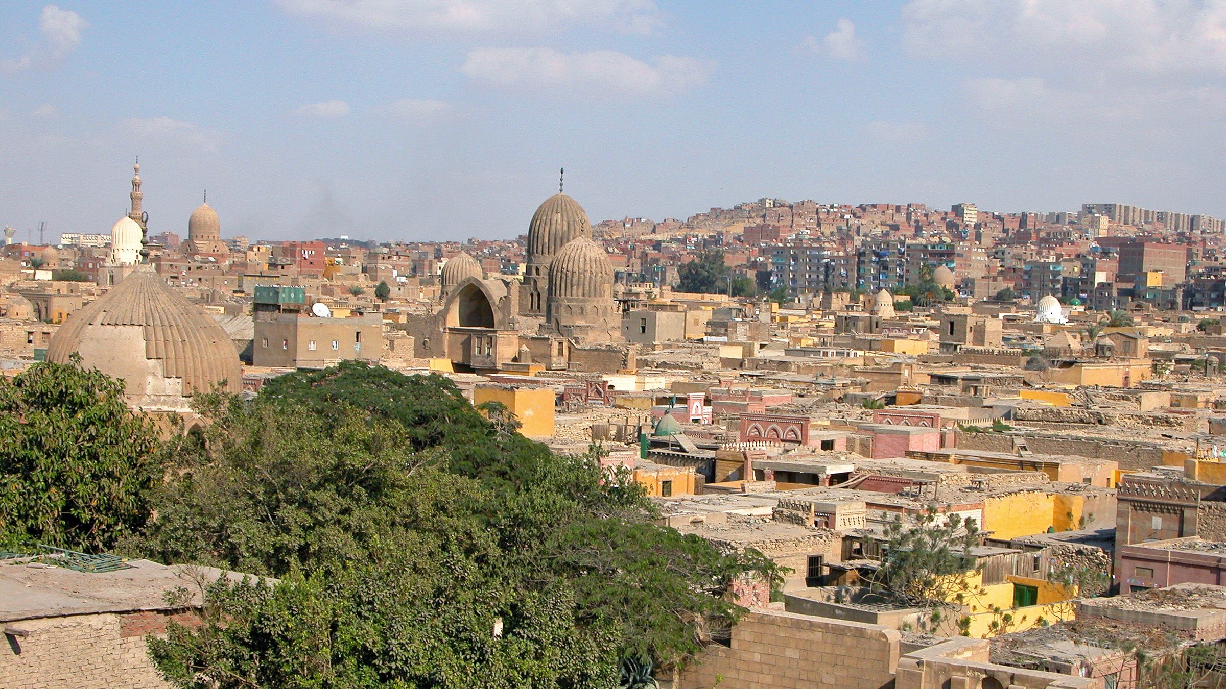L'image montre une vue panoramique d'une ville historique, probablement un quartier ancien avec des bâtiments en pierre. On peut voir plusieurs coupoles et minarets qui témoignent de l'architecture traditionnelle. Les toits sont variés, certains sont plats et d'autres sont plus élevés, tandis que la végétation semble présente avec des arbres. En arrière-plan, on aperçoit des collines et d'autres bâtiments, suggérant une ville vaste et densément peuplée. Le ciel est clair avec quelques nuages, donnant une atmosphère lumineuse à la scène.
