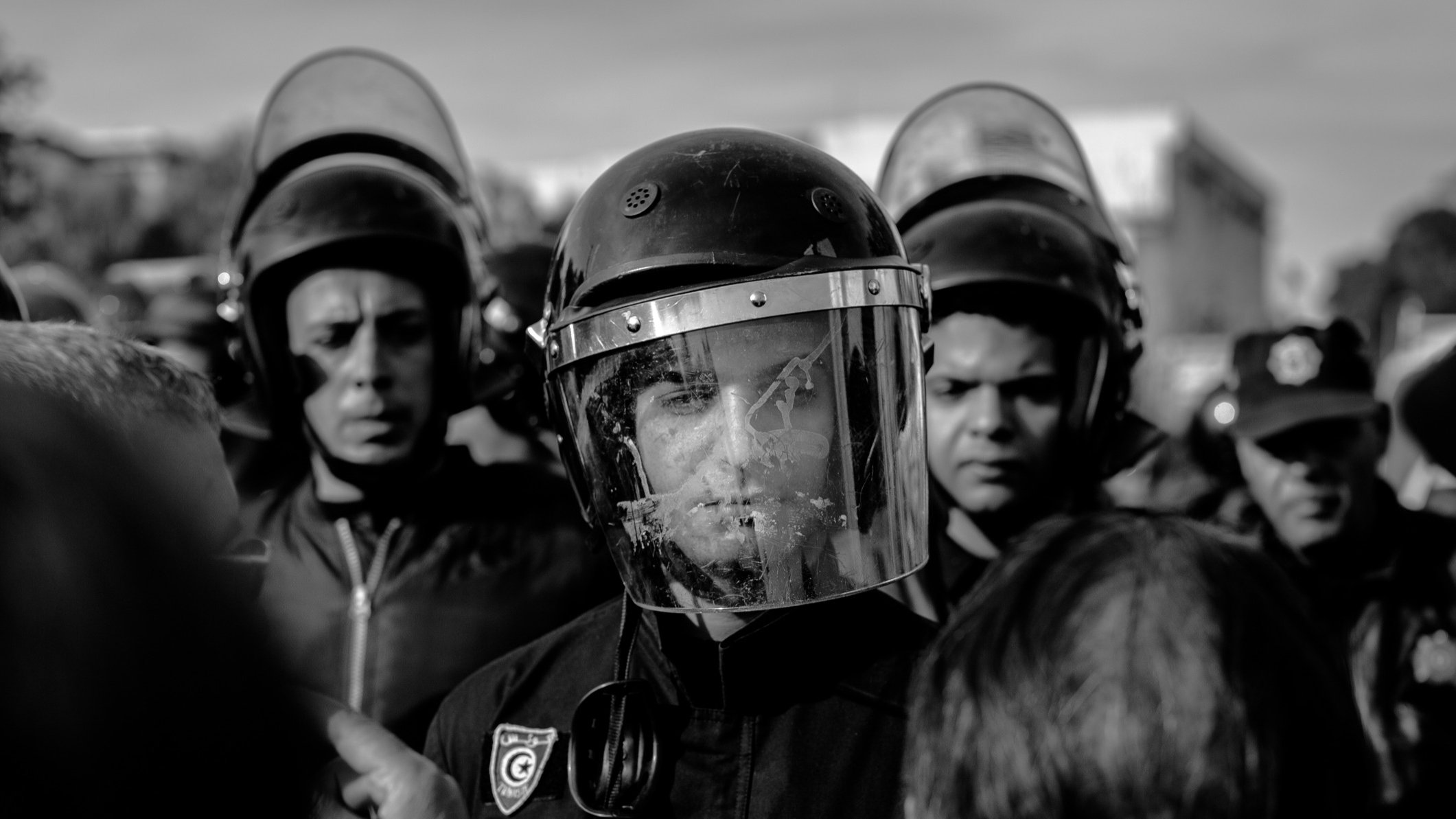 L'image montre un groupe de personnes, probablement des agents des forces de l'ordre, entourés par une foule. L'un des agents est au premier plan, portant un casque et un bouclier facial, ce qui indique qu'il est en position de maintien de l'ordre. Les visages des autres personnes en arrière-plan sont également visibles, exprimant une certaine tension. L'image est en noir et blanc, ce qui accentue le sérieux de la situation. L'atmosphère semble chargée, avec un sentiment de confrontation possible.