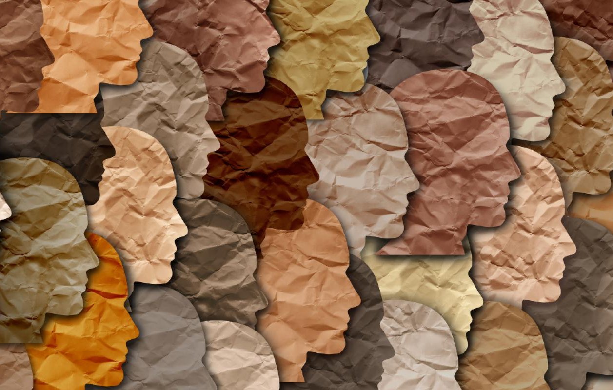 The image features a collage of human profiles created from various shades of crumpled paper. Each silhouette has a different tone, representing a diverse range of skin colors. The overlapping and varied textures of the paper give depth and interest to the composition, highlighting themes of diversity and inclusion. The overall effect is a harmonious blend of shapes and colors.