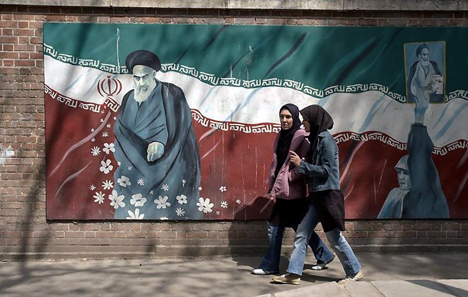 الصورة تظهر جدارية على جدار مبني، حيث تبرز شخصيتان متجولتان في الشارع. الجدارية تحتوي على رسم لقائد تاريخي إيراني، مع العلم الإيراني في الخلفية. الفتيات اللاتي يمررن ترتدين الحجاب، ويبدو أنهن في وضع محادثة. تتضمن الجدارية أيضاً بعض الزهور، مما يضفي لمسة جمالية على المشهد.