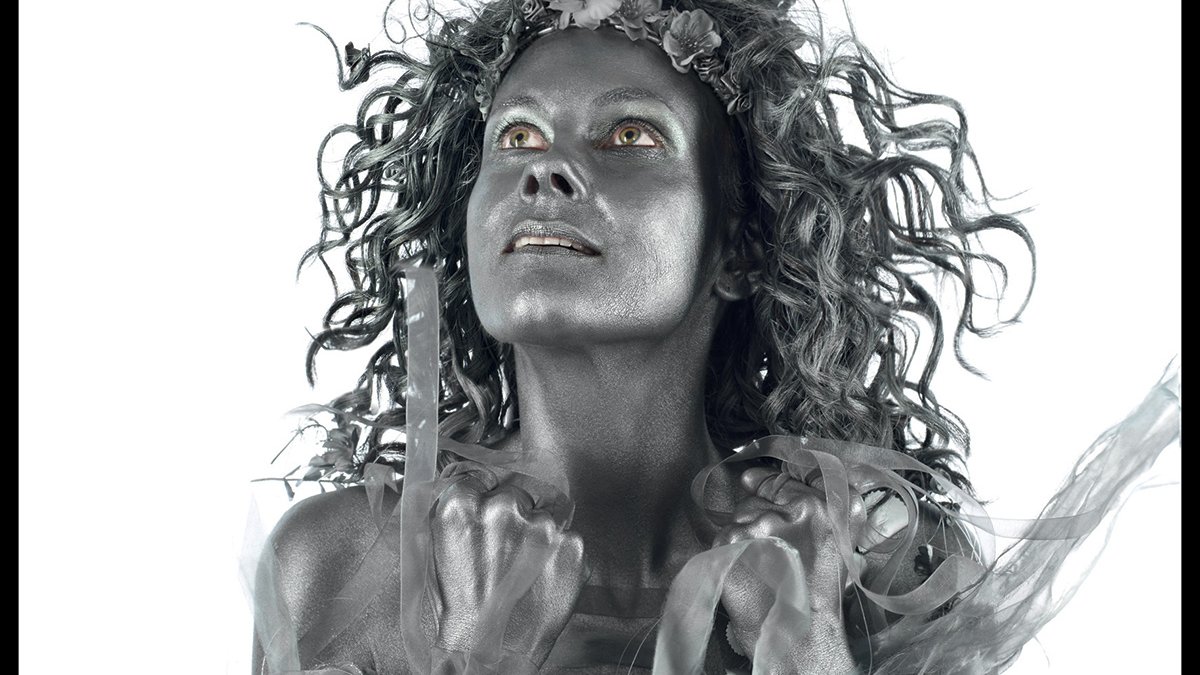 L'image affiche une femme peinte en argent, avec des cheveux ébouriffés et un regard intense. Elle est présentée dans un style dramatique, évoquant une performance artistique. Le fond est noir, ce qui met en avant la couleur métallique de son corps. En haut, on voit le titre de l'événement "D(rôles de printemps)" en grandes lettres, suivi des informations sur le lieu et les dates de l'événement, qui se déroule au TARMAC à Paris. Des détails supplémentaires sur les performances de théâtre et de danse sont également mentionnés, soulignant la diversité culturelle et artistique de l'événement.