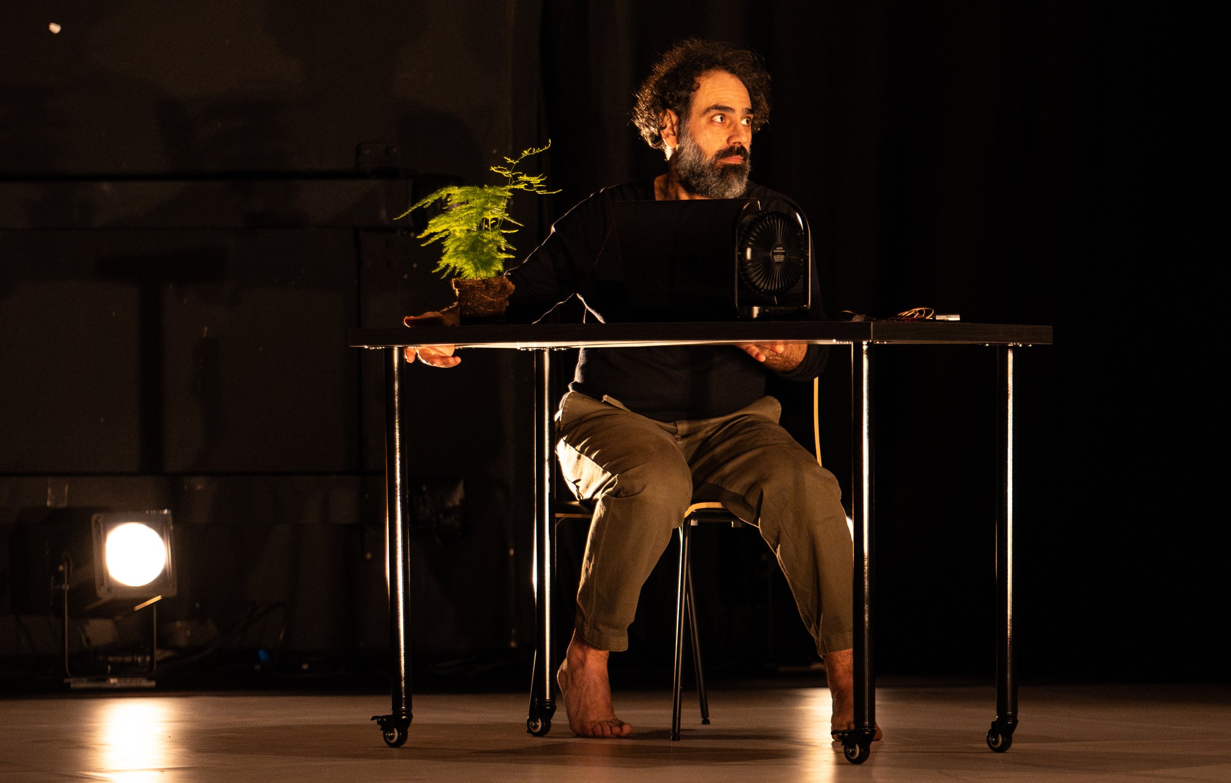 L'immagine mostra un uomo seduto a un tavolo in un ambiente di scena illuminato. Indossa una maglietta nera e pantaloni chiari, e sembra concentrato su un oggetto davanti a lui. Accanto a lui c'è una piccola pianta in vaso, mentre una ventola è posizionata sulla sua sinistra. L'illuminazione crea un'atmosfera drammatica, evidenziando sia l'uomo che gli oggetti sul tavolo. L'ambiente è scuro, con alcune luci che si riflettono sul pavimento.