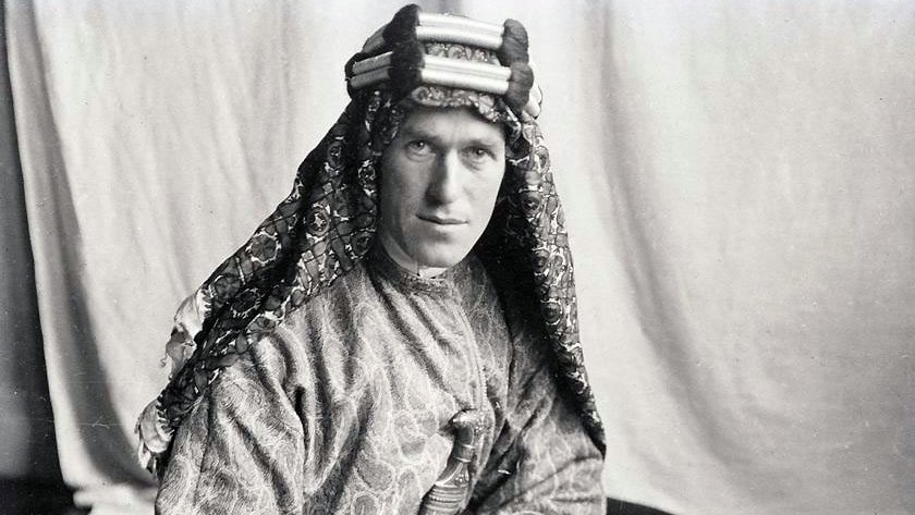 L'image montre un homme portant un costume traditionnel. Il est vêtu d'une tunique ornée et d'un headdress décoratif. Son regard est sérieux et il tient un livre dans ses mains. L'arrière-plan est sobre, ce qui met en valeur son apparence et son habillement. La photo semble être ancienne, ce qui lui donne un aspect historique.