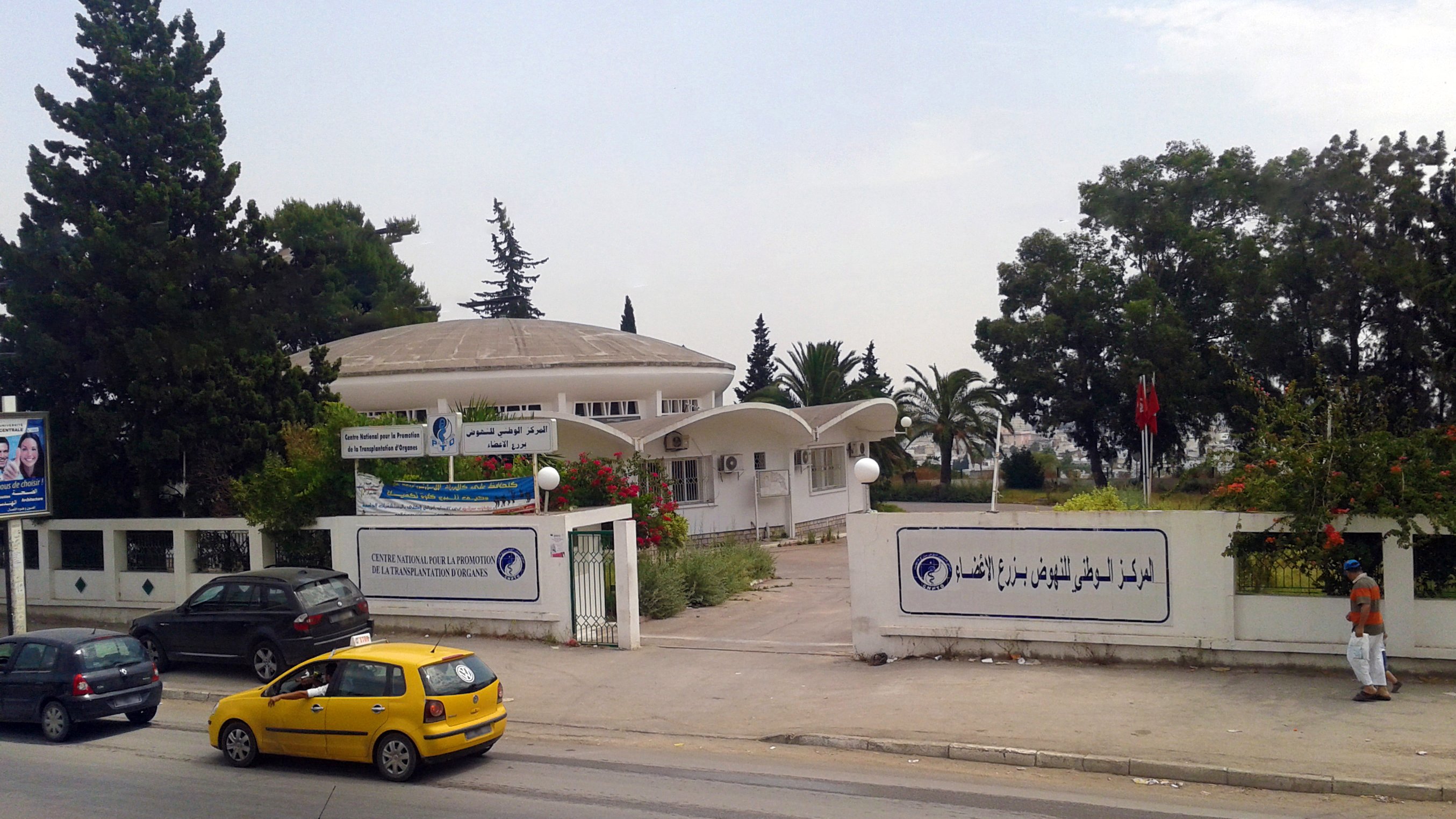 L'image montre un bâtiment moderne avec un toit arrondi, entouré de verdure et d'arbres. On peut voir une clôture blanche et plusieurs panneaux affichant des informations. À l'avant, des véhicules sont garés le long de la route, dont une voiture jaune. L'environnement paraît calme, et le ciel est dégagé, ce qui donne une impression de tranquillité.