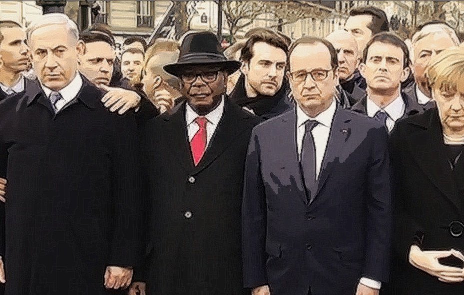 L'image montre un groupe de personnes en émoi, rassemblées dans un cadre public, probablement lors d'un événement solennel. On peut y voir des figures politiques en costumes, dont certaines portent des démarches sérieuses et concentrées. L'ambiance semble être empreinte de gravité et de solidarité. Les participants sont alignés et se regardent, ce qui indique une atmosphère de recueillement ou de soutien mutuel.