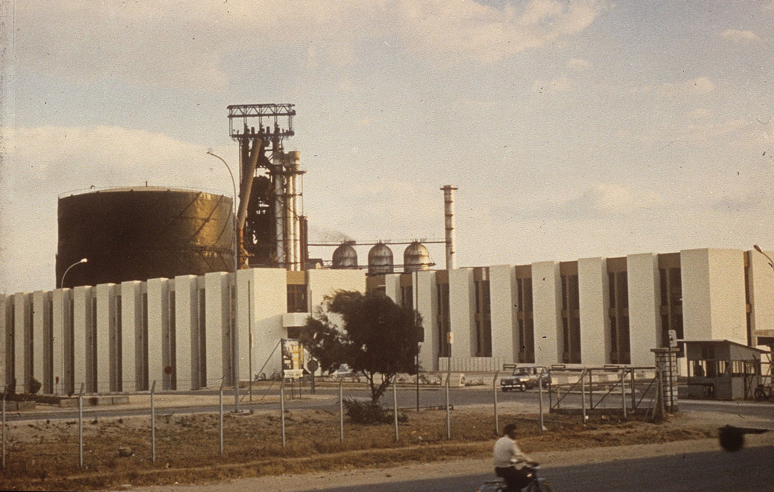 L'image montre une grande installation industrielle. On peut voir un grand réservoir noir et une structure avec des cheminées et des tubes. La façade de l'immeuble est faite de blocs blancs. À l'avant, il y a une route, et on aperçoit une personne à vélo sur celle-ci. L'environnement semble être un site industriel, avec quelques arbres et une clôture qui délimite la zone. Le ciel est partiellement nuageux, ce qui donne une ambiance légèrement vintage à la scène.