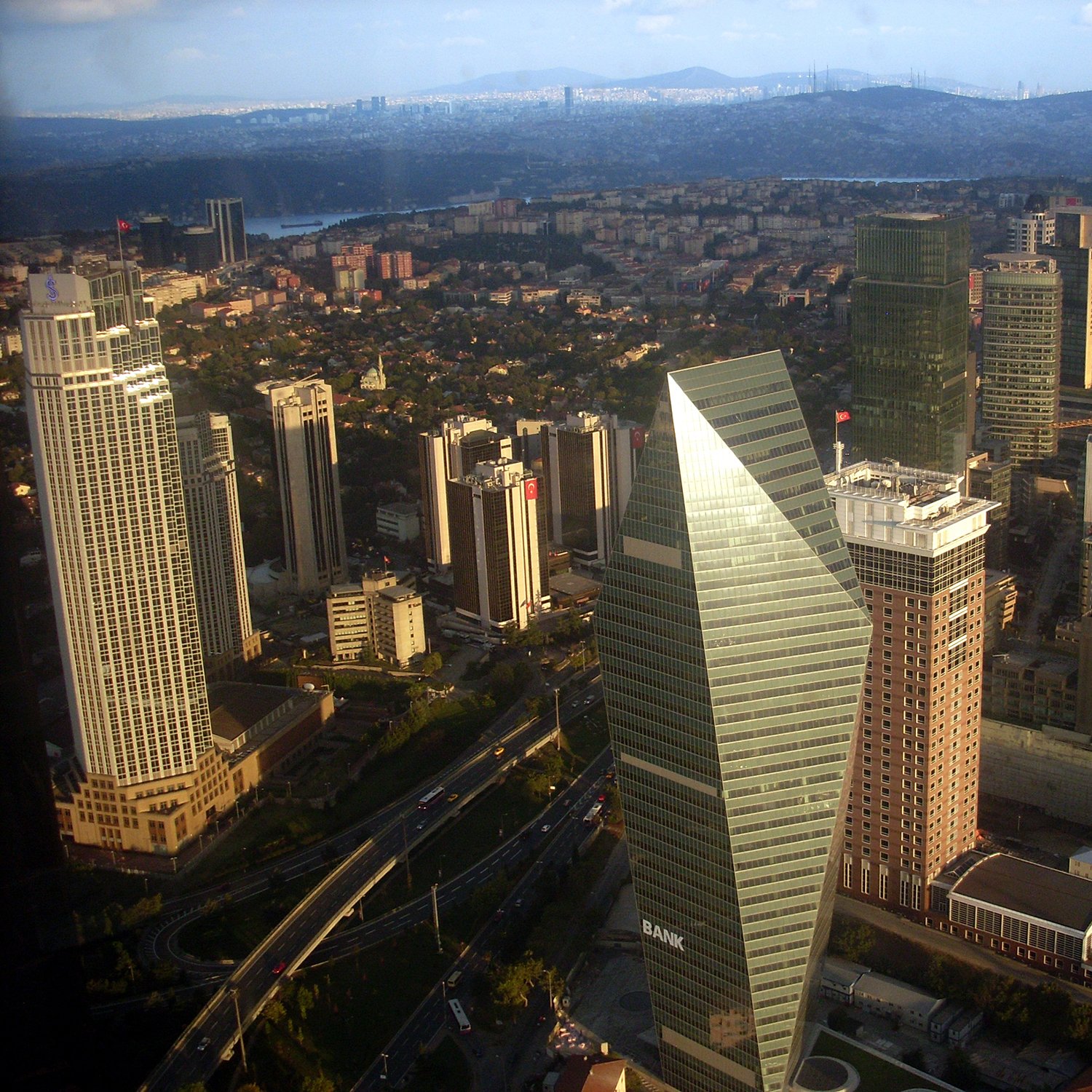 این تصویر نمایی از یک شهر مدرن است که در آن ساختمان‌های بلند و شیک مشاهده می‌شوند. در مرکز تصویر یک ساختمان با طراحی متمایز و زاویه‌دار وجود دارد که به‌نظر می‌رسد نماد یکی از بانک‌ها باشد. دیگر ساختمان‌ها نیز به ارتفاع و تنوع طراحی در اطراف آن قرار دارند. فضای سبز و خیابان‌های در حال تردد نیز در بخش‌های پایین تصویر قابل مشاهده است. آسمان آبی با ابرهای خفیف به زیبایی تصویر افزوده است.