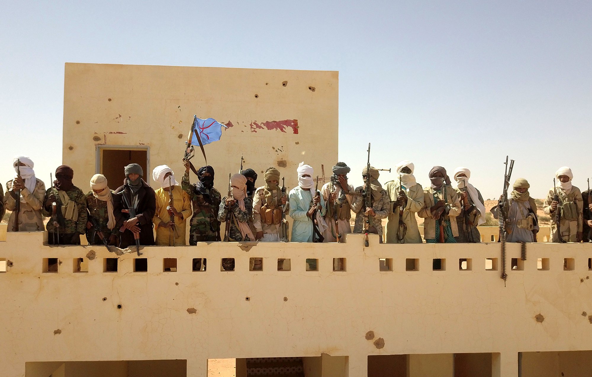 L'image montre un groupe de personnes armées, probablement des membres d'un groupe militant, se tenant sur un balcon ou une terrasse d'un bâtiment. Ils portent des vêtements traditionnels et des masques, et certains brandissent des armes. Le fond est désertique et ensoleillé, avec un ciel clair. On remarque également un drapeau dans le groupe, suggérant un certain degré d'organisation ou d'identité collective.