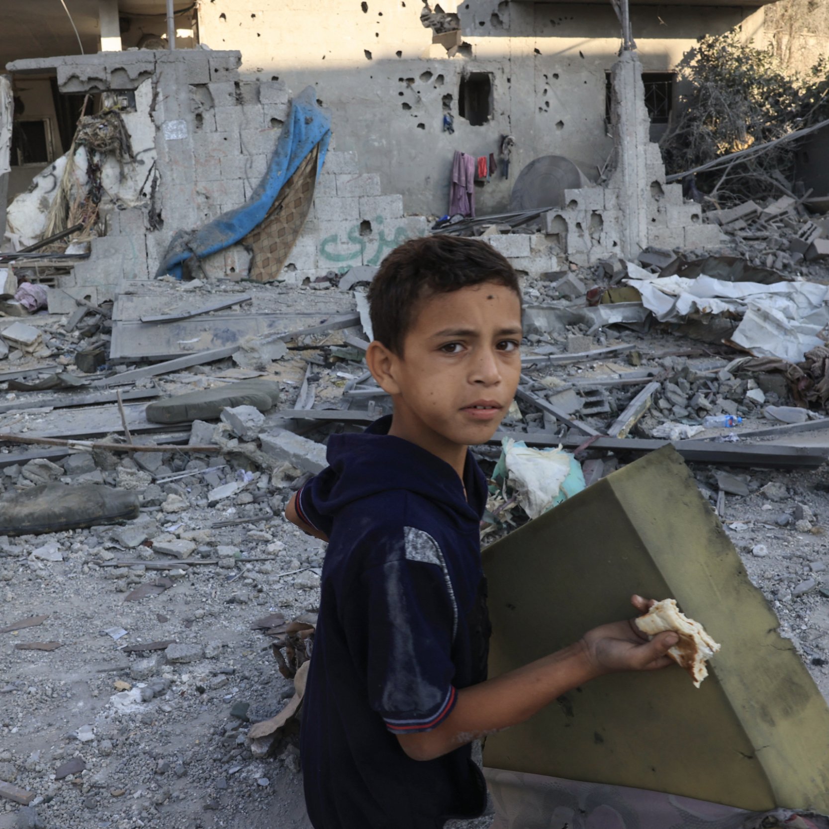 L'image montre un jeune garçon au visage marqué par l'environnement chaotique autour de lui. Il porte des vêtements simples et tient un morceau qu'il semble avoir trouvé dans les débris. Le fond est constitué de bâtiments détruits, avec des murs endommagés et des débris éparpillés, ce qui évoque une situation de conflit ou de catastrophe. La lumière du jour éclaire la scène, mettant en évidence la gravité de la situation dans laquelle il se trouve.
