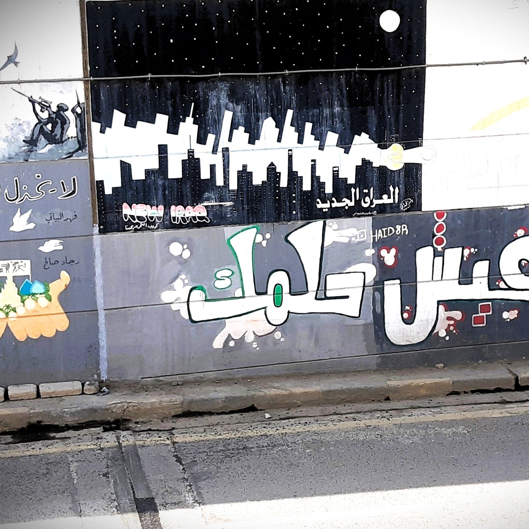 L'immagine mostra un murale colorato su un muro, con diversi disegni e scritte in arabo. In primo piano, si legge "عيش حلمك" che significa "Vivi il tuo sogno". Lo sfondo presenta vari elementi artistici, tra cui silhouette di grattacieli e figure stilizzate, creando un'atmosfera urbana e vibrante. Ci sono anche elementi decorativi e simboli che riflettono un messaggio di speranza e ispirazione.