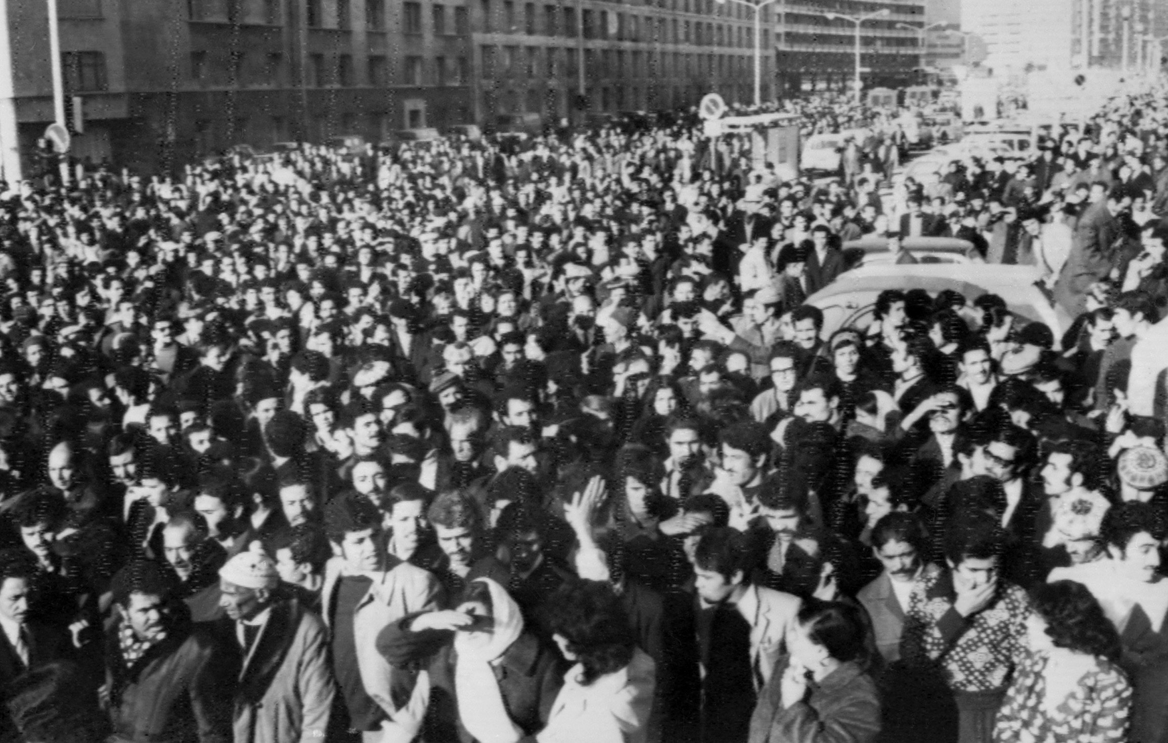 The image depicts a large crowd of people gathered in an urban setting, likely during a significant event or protest. The crowd appears to be densely packed, filling the street and surrounding areas. Many individuals are dressed in formal or semi-formal attire, indicating the seriousness of the occasion. The atmosphere seems charged, reflecting a sense of unity and purpose among the participants. Buildings line the background, and there are vehicles partially visible, suggesting a city environment. The black-and-white effect adds a historical or dramatic tone to the scene.