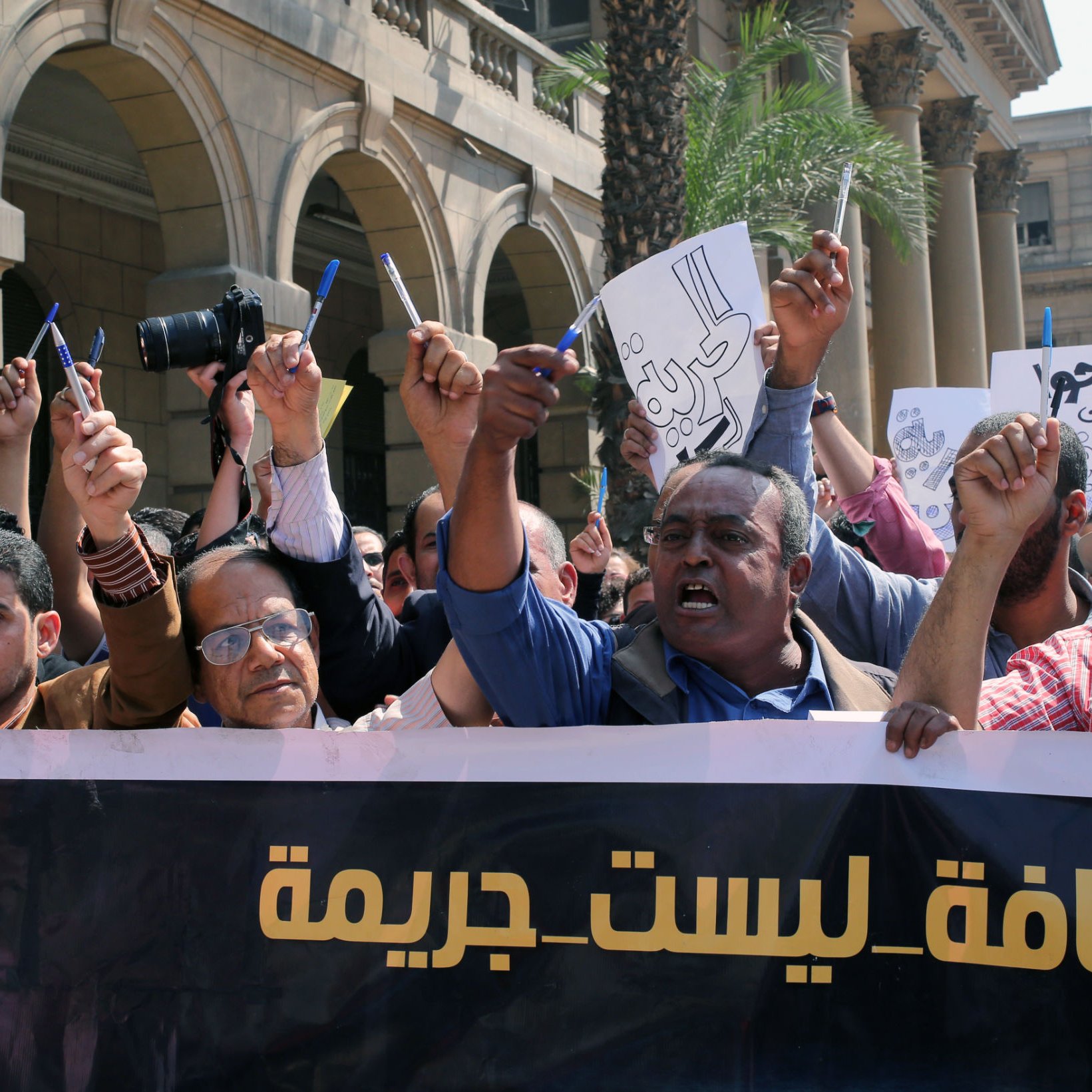 تظهر في الصورة مظاهرة تضم مجموعة من الأشخاص يحملون لافتات وأعلامًا. يرفع البعض أيديهم بشكل تعبيري، بينما يبدو البعض الآخر يتحدث أو يحتج. يوجد شعارات على اللافتات تعبر عن قضايا تتعلق بالصحافة وحرية التعبير، حيث يظهر نص مكتوب يشير إلى أن الصحافة ليست جريمة. الخلفية تحتوي على مباني كبيرة، مما يشير إلى أن المظاهرة قد تكون حدثت في مكان عام أو أمام منشأة حكومية.