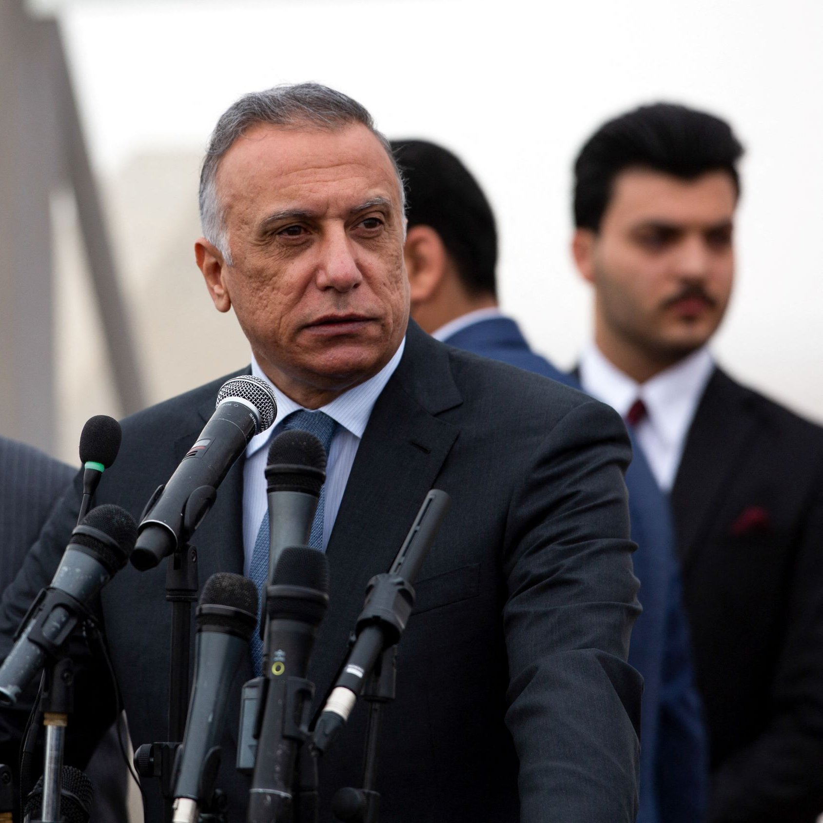 در این تصویر، دو فرد در یک کنفرانس خبری یا مراسم رسمی دیده می‌شوند. فردی که در سمت راست قرار دارد، لباس رسمی پوشیده و به نظر می‌رسد در حال پاسخ دادن به سوالات یا صحبت کردن است. او چند میکروفن مقابل خود دارد که نشان از اهمیت موضوع دارد. فرد دیگر در سمت چپ نیز لباس رسمی دارد و در حال گوش دادن به صحبت‌های فرد اول است. پس‌زمینه تصویر کمی مبهم به نظر می‌رسد. وضوح و جزئیات چهره‌ها به دلیل زاویه و ترکیب تصویر محدود است.