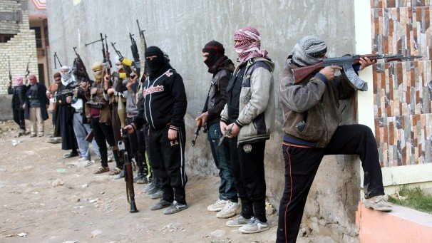 L'image montre un groupe de personnes armées, se tenant alignées le long d'un mur. Elles portent des vêtements sombres et des couvertures sur la tête, ce qui masque leurs visages. Chacune d'elles tient une arme à feu, et certaines adoptent des postures prêtes au combat. L'environnement semble urbain, avec des bâtiments en arrière-plan et un terrain sale. L'atmosphère générale est tendue et évoque un contexte de conflit ou de violence.