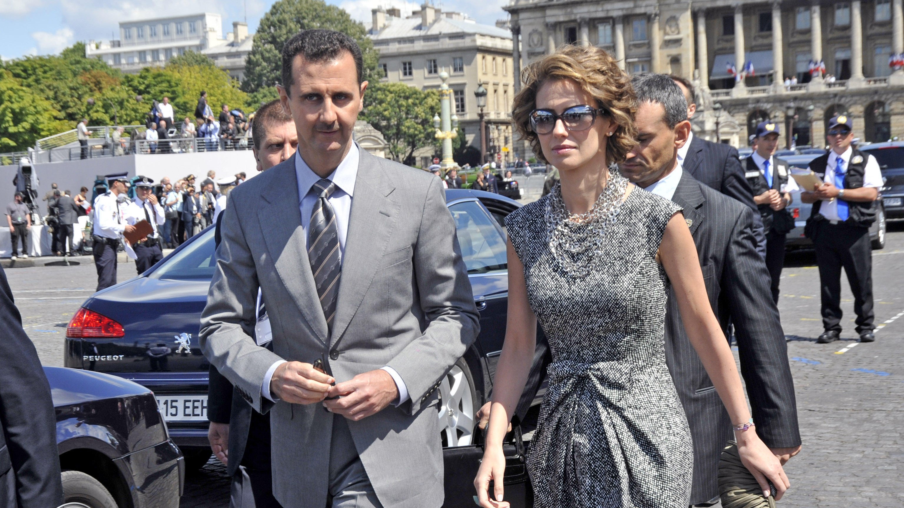 The image shows a man and a woman walking together in an outdoor setting. The man is dressed in a grey suit and tie, while the woman is wearing a stylish, patterned dress, sunglasses, and holds a handbag. They appear to be in a public area, possibly during a formal event, with a group of people in the background, including security personnel. The architecture behind them suggests a significant building or landmark.