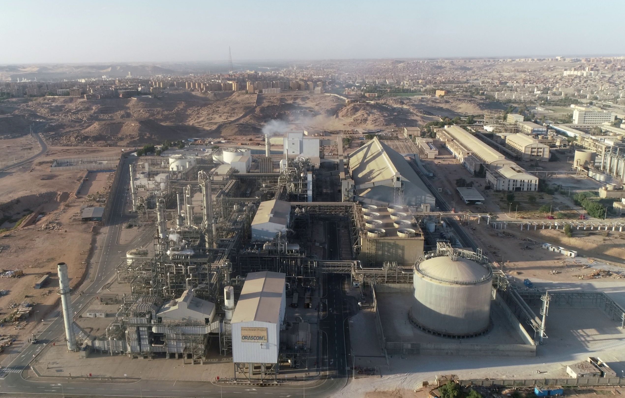 L'image présente une vue aérienne d'un complexe industriel. On peut observer plusieurs bâtiments de grande taille, comprenant des structures cylindriques et pyramidales. La zone environnante semble aride avec des collines et des bâtiments à l'arrière-plan. Des tuyaux et des infrastructures industrielles sont visibles, indiquant une activité de production. On discerne également de la vapeur s'échappant d'une des installations, suggérant un processus en cours. L'ensemble dénote un environnement industriel dans une région désertique.