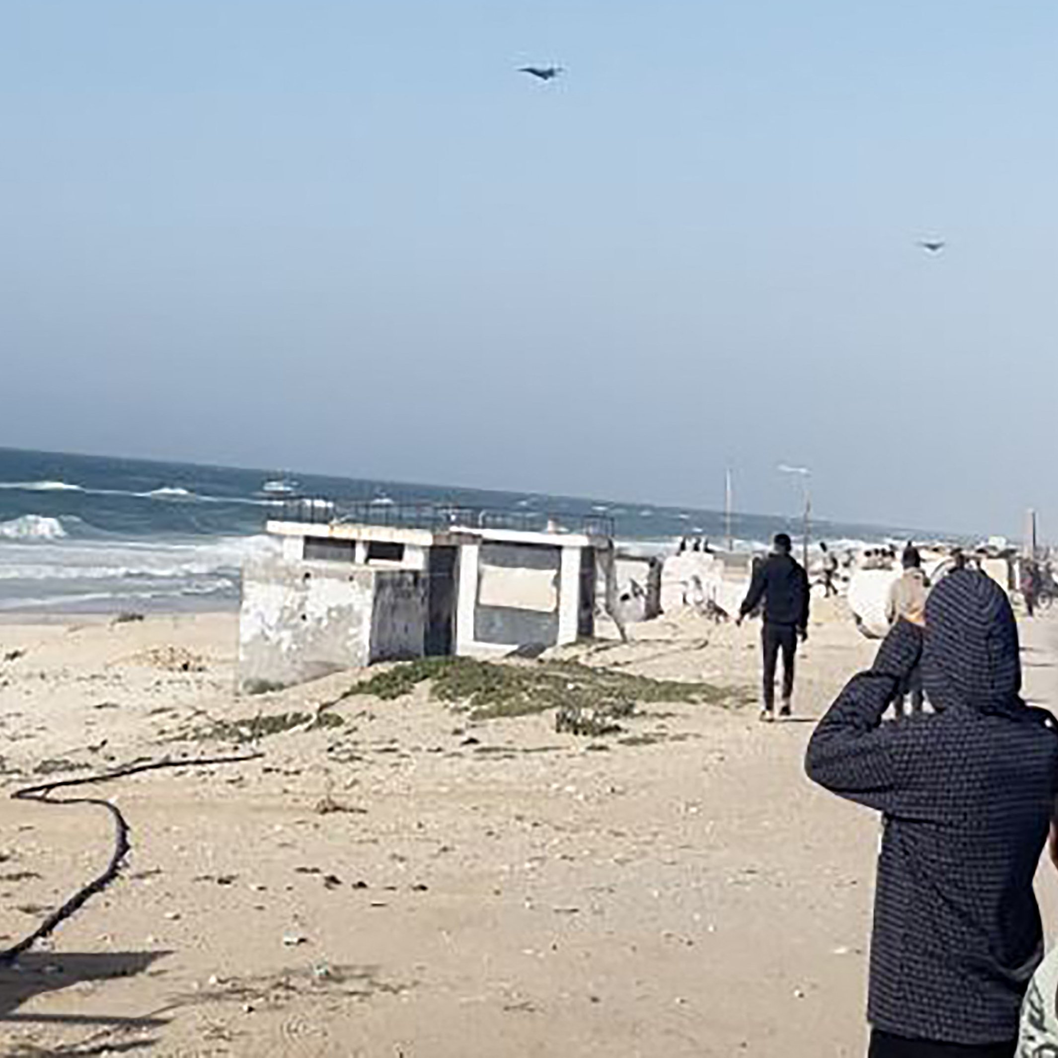 L'immagine mostra una scena costiera con il mare e delle onde in movimento. Si possono vedere alcune persone sulla spiaggia che osservano il mare, apparentemente interessate a qualcosa in cielo. Il paesaggio è caratterizzato da sabbia chiara e una scalinata che porta a una struttura, mentre in lontananza si vedono veicoli e persone che si muovono lungo la costa. Il cielo è sereno, con poche nuvole.