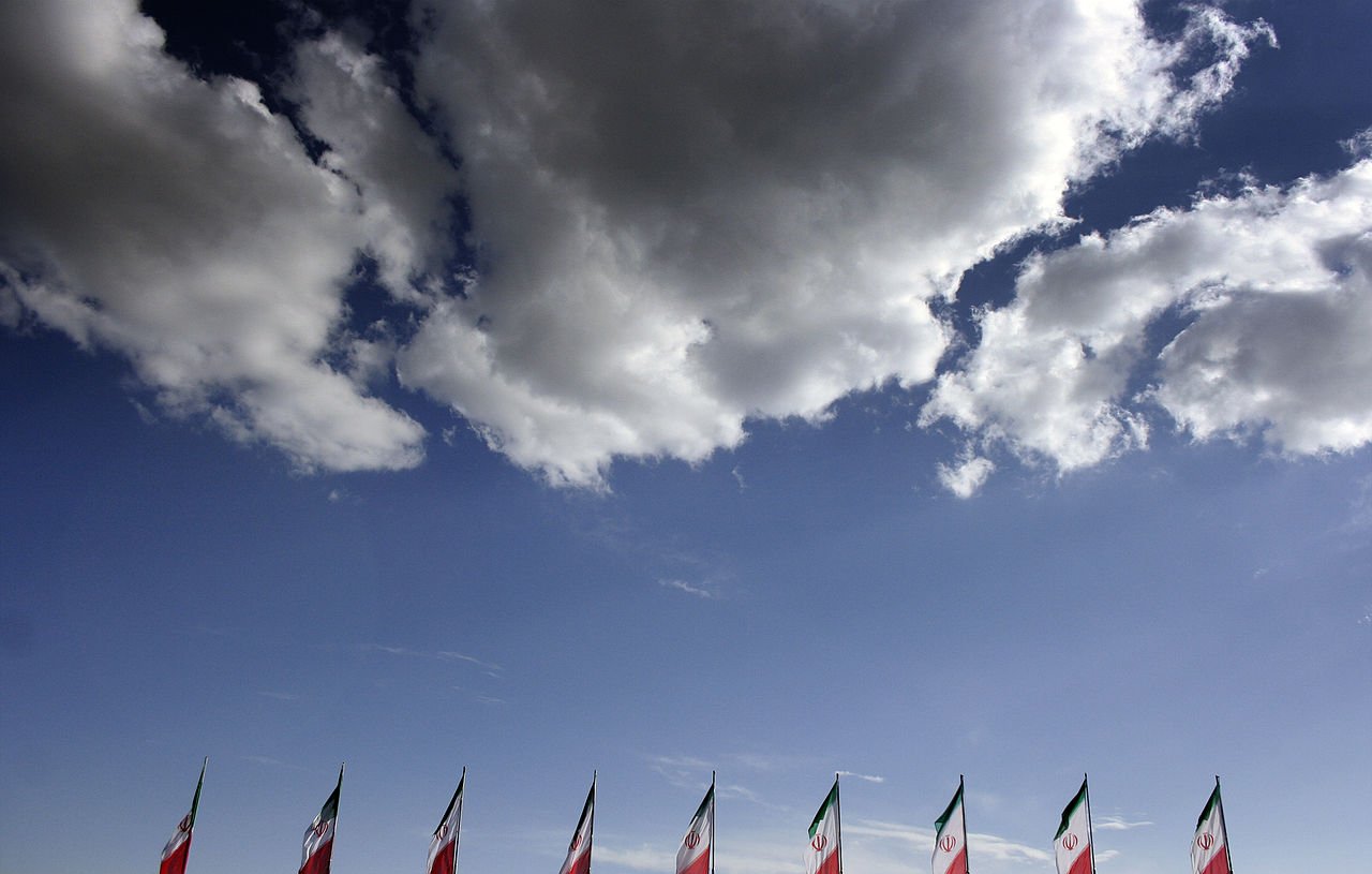 در این تصویر، چند پرچم در حال اهتزاز در برابر آسمانی آبی و ابری دیده می‌شود. پرچم‌ها به رنگ‌های سبز، سفید و قرمز هستند که رنگ‌های ملی ایران را نمایندگی می‌کنند. ابرهای سفید نیز در پس‌زمینه به زیبایی آسمان را تزئین کرده‌اند. این ترکیب تصویری از یک روز آفتابی و شاداب را به نمایش می‌گذارد.