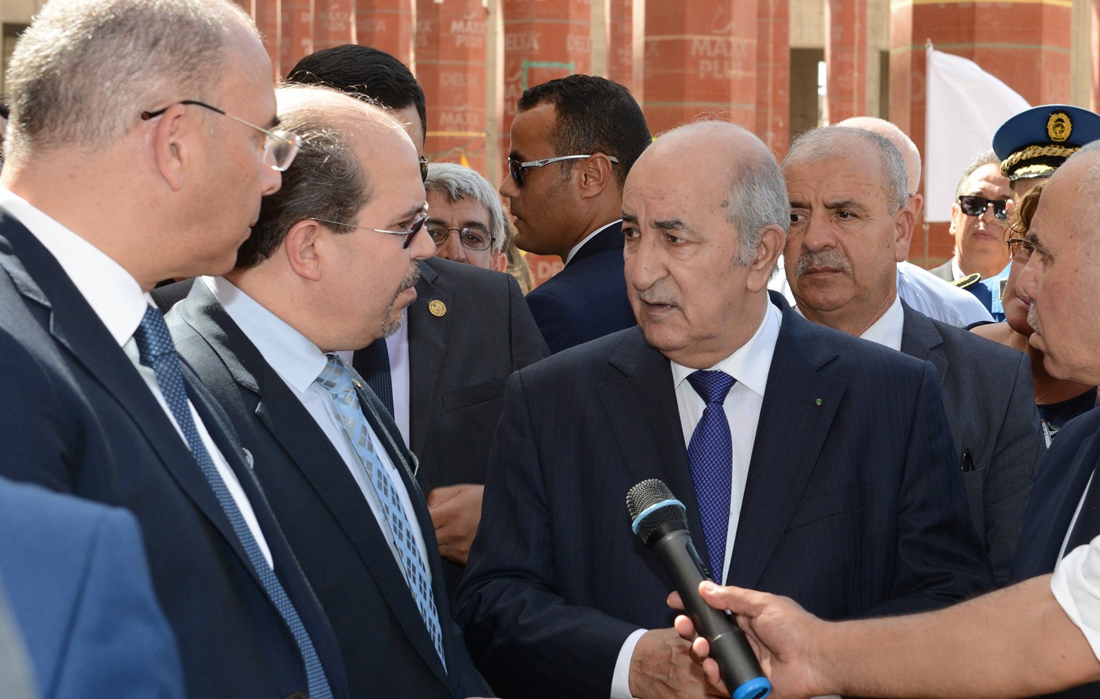 L'image montre un groupe d'hommes en costume, probablement en train de discuter. Au centre, l'un des hommes semble occuper une position de leader, tandis que les autres l'écoutent attentivement. Ils semblent être dans un cadre formel, peut-être lors d'une visite officielle ou d'un événement important. L'environnement en arrière-plan suggère un lieu institutionnel, avec des structures architecturales visibles.