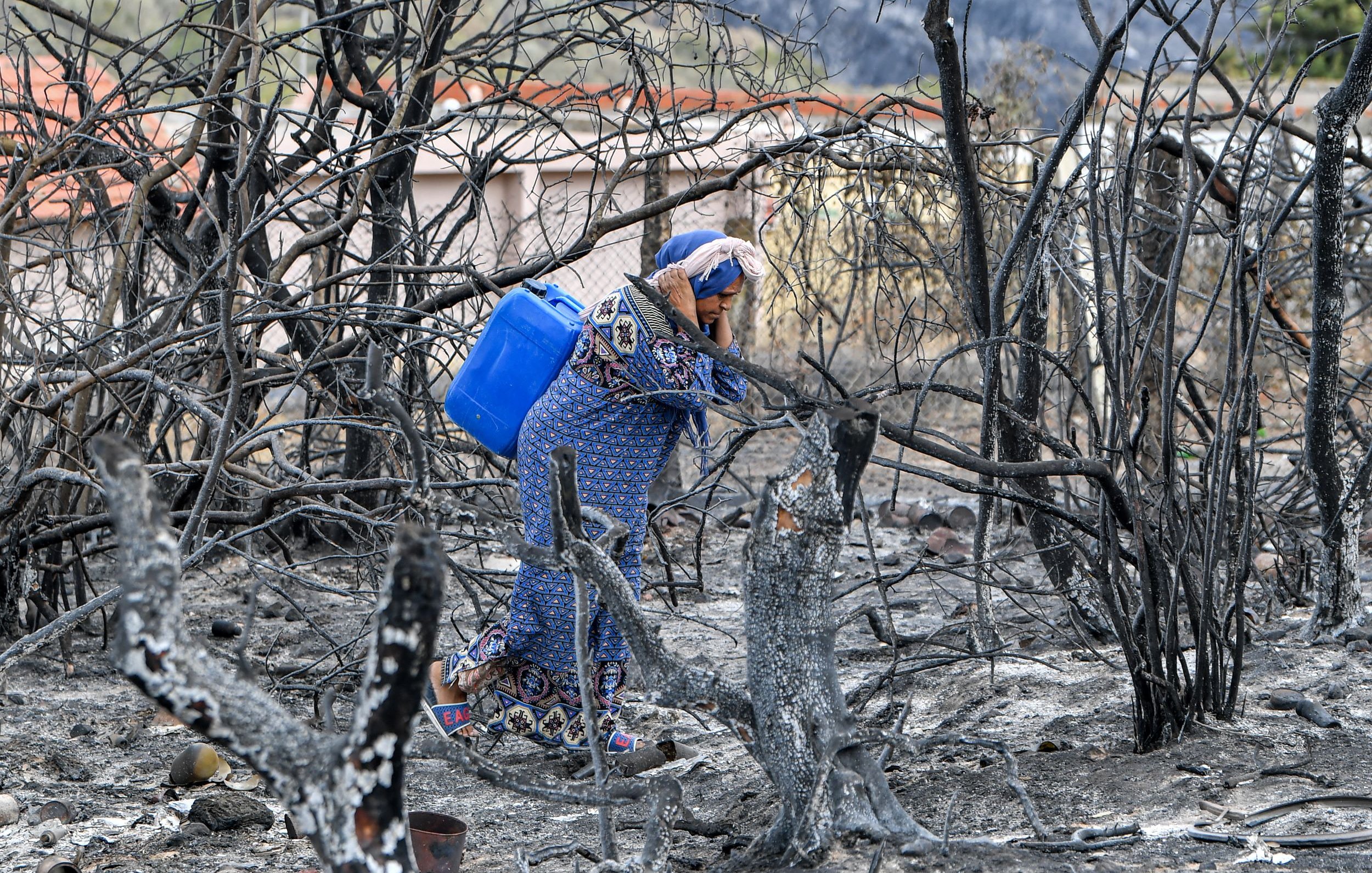 The image depicts a woman wearing a patterned dress and headscarf, walking through a landscape that has been ravaged by fire. The ground is charred and blackened, with the remnants of burned trees and vegetation surrounding her. She is carrying a blue container, possibly for water, as she navigates through the bleak, desolate environment. The backdrop includes hints of buildings, suggesting that this area has been impacted by a wildfire. The scene conveys a sense of loss and resilience amidst the devastation.