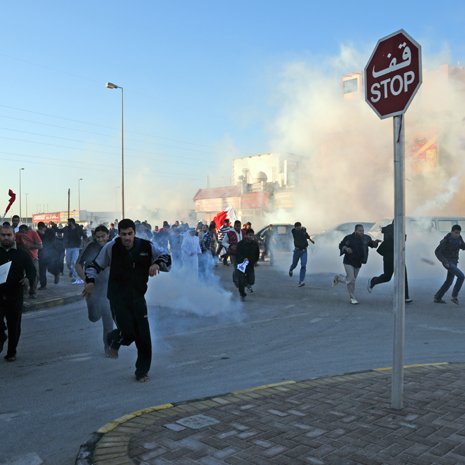 L'image montre une scène de manifestation. De nombreuses personnes courent dans une rue, entourées de fumée, probablement due à des gaz lacrymogènes. Au premier plan, on peut voir un panneau de signalisation "STOP". L'atmosphère semble tendue, et les manifestants semblent fuir une situation chaotique. Les participants portent des vêtements variés et certains tiennent des drapeaux.