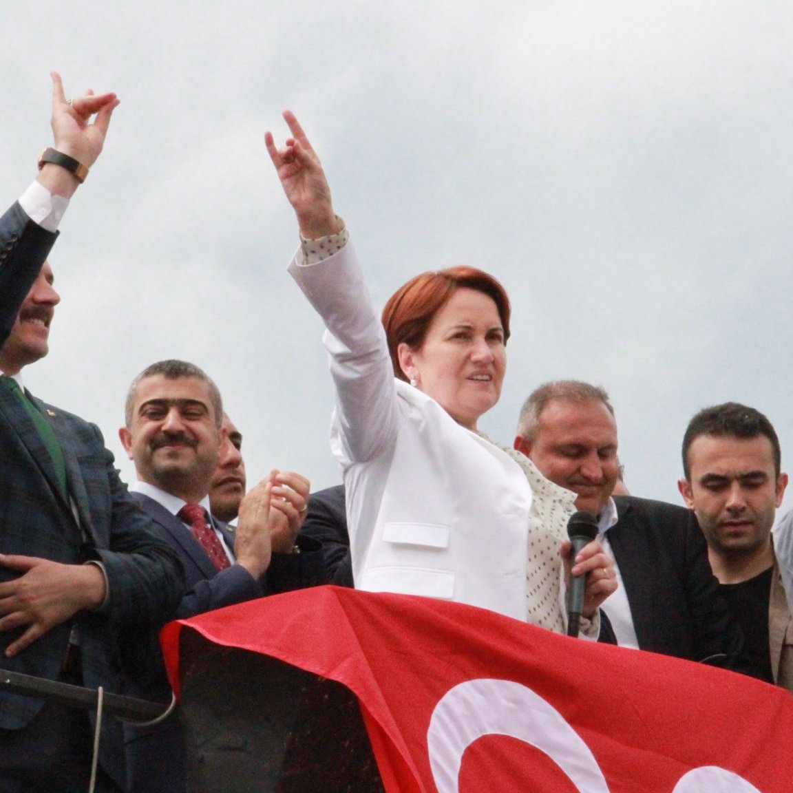 The image depicts a group of people at a political rally or event. In the foreground, a woman dressed in a white suit is speaking into a microphone, raising her arm with a gesture that signifies support or determination. Behind her, several men are visible, some clapping and others making similar gestures. The background features a large red banner, which may be associated with the event or political party. The overall atmosphere seems to convey enthusiasm and solidarity among the attendees.