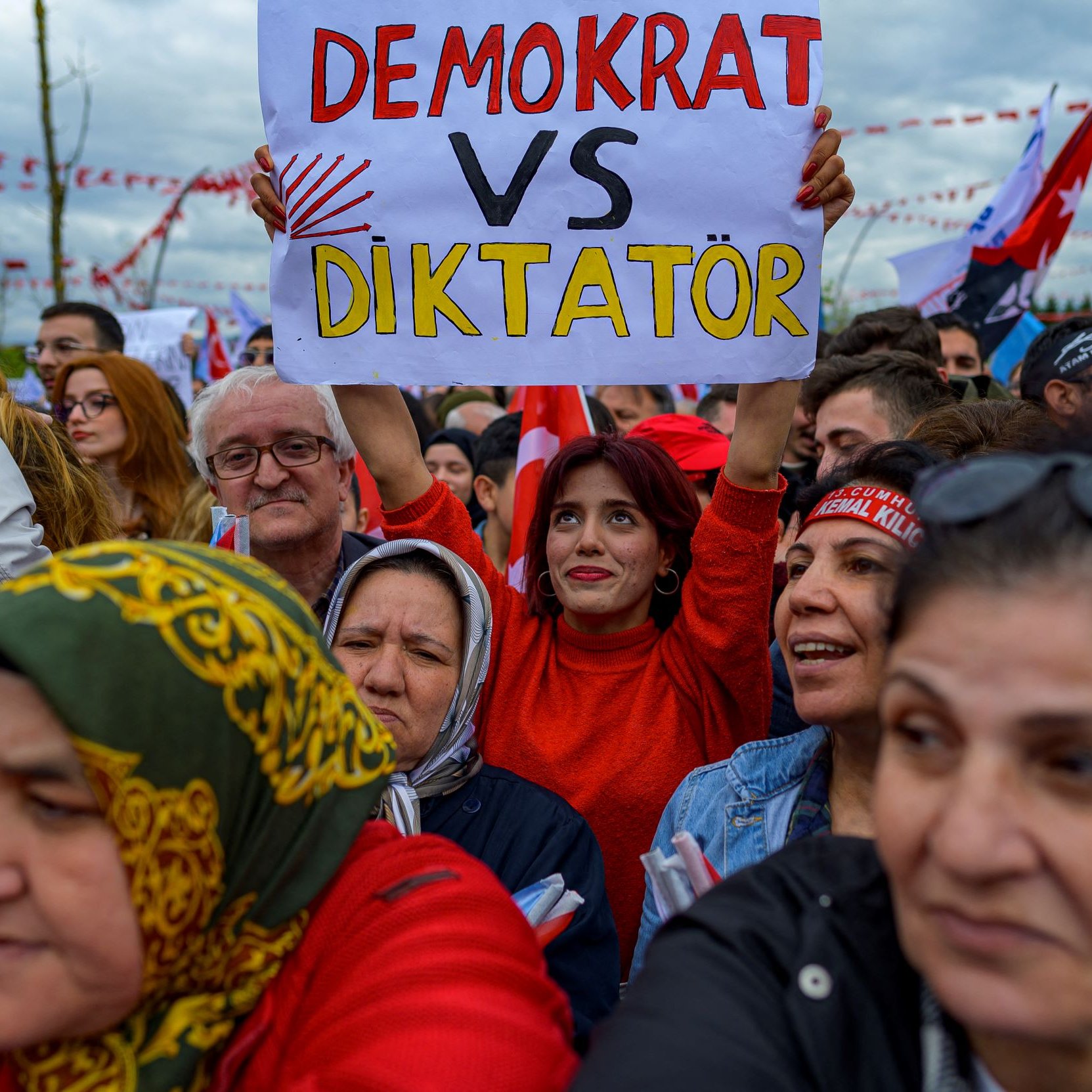 Nell'immagine si vede un gruppo di persone ad una manifestazione. In primo piano, una donna sorridente indossa una maglietta rossa e tiene in mano un cartello con la scritta "DEMOCRAT VS DIKTATÖR". Intorno a lei, ci sono diverse altre persone che sembrano partecipare attivamente all'evento. Molti di loro indossano sciarpe e cappelli colorati. Sullo sfondo, si vedono bandiere e decorazioni che contribuiscono all'atmosfera di protesta. L'ambientazione sembra carica di emozioni e impegno civico.