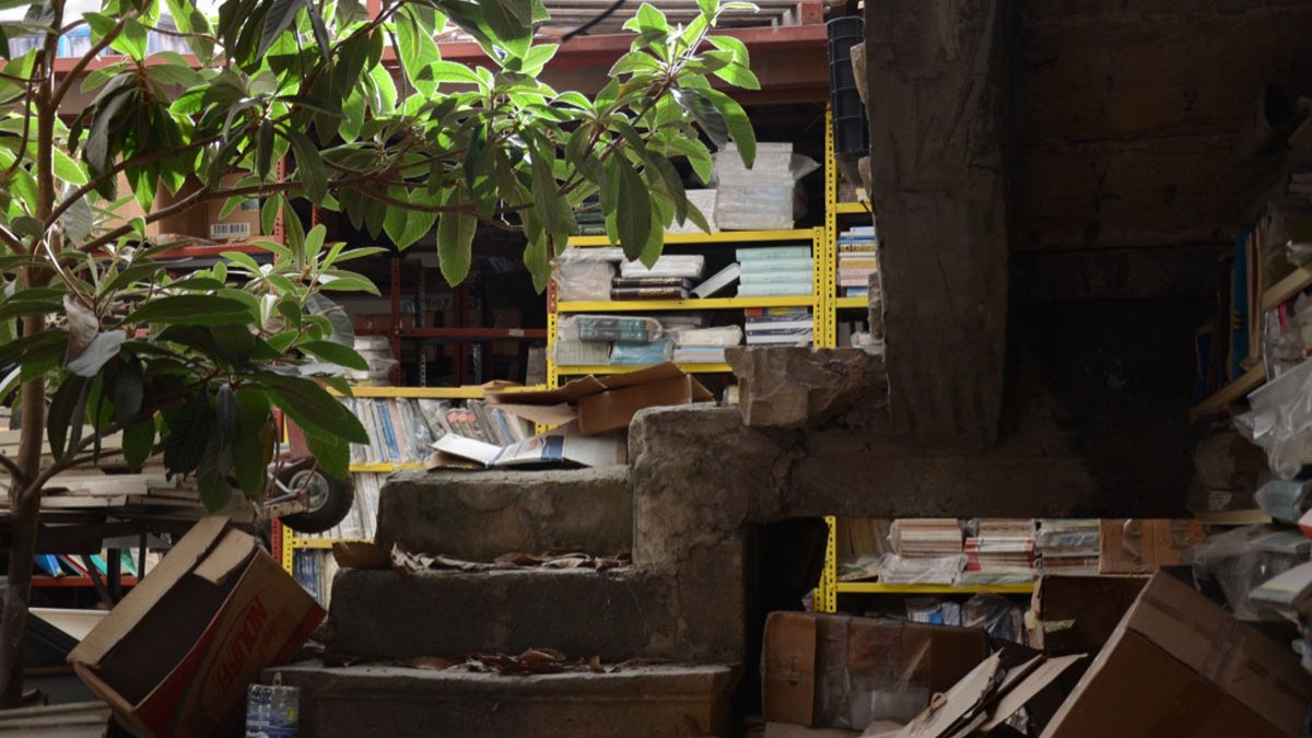 L'image montre un espace intérieur, apparemment désordonné. On y voit un escalier en béton partiellement recouvert de débris et de boîtes. À côté de l'escalier, il y a une plante verte, ce qui ajoute une touche de vie à cet environnement. En arrière-plan, des étagères remplies de livres et d'objets se pressent, donnant l'impression que l'endroit est utilisé comme entrepôt ou bibliothèque abandonnée. Les lumières naturelles entrent par une ouverture en haut, éclairant partiellement la scène.
