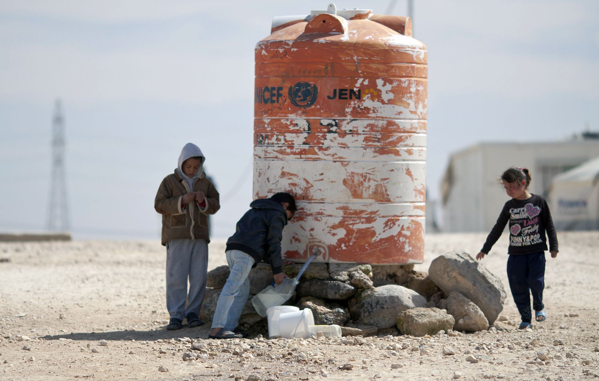تظهر الصورة مجموعة من الأطفال بالقرب من خزان ماء كبير مُسجّل عليه علامة الأمم المتحدة. يقف اثنان من الأطفال بجانب الخزان، حيث يبدو أحدهم مشغولاً في فحص شيء ما، بينما يقوم الآخر بسكب الماء في وعاء. هناك طفل ثالث يلعب بجانبهم. الخلفية تبدو قاحلة مع بعض الصخور والأرض الرملية، مما يشير إلى بيئة جافة.