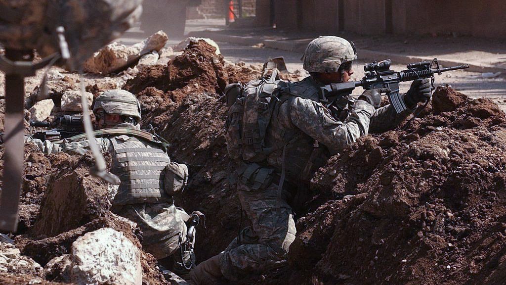 L'image montre des soldats en engagement militaire, se cachant derrière des tranchées. Ils portent des uniformes militaires et des casques, et sont armés. Le terrain est marqué par des gravats et de la terre, suggérant un environnement de combat. L'atmosphère est tendue, montrant la préparation et la vigilance des soldats face à un danger.
