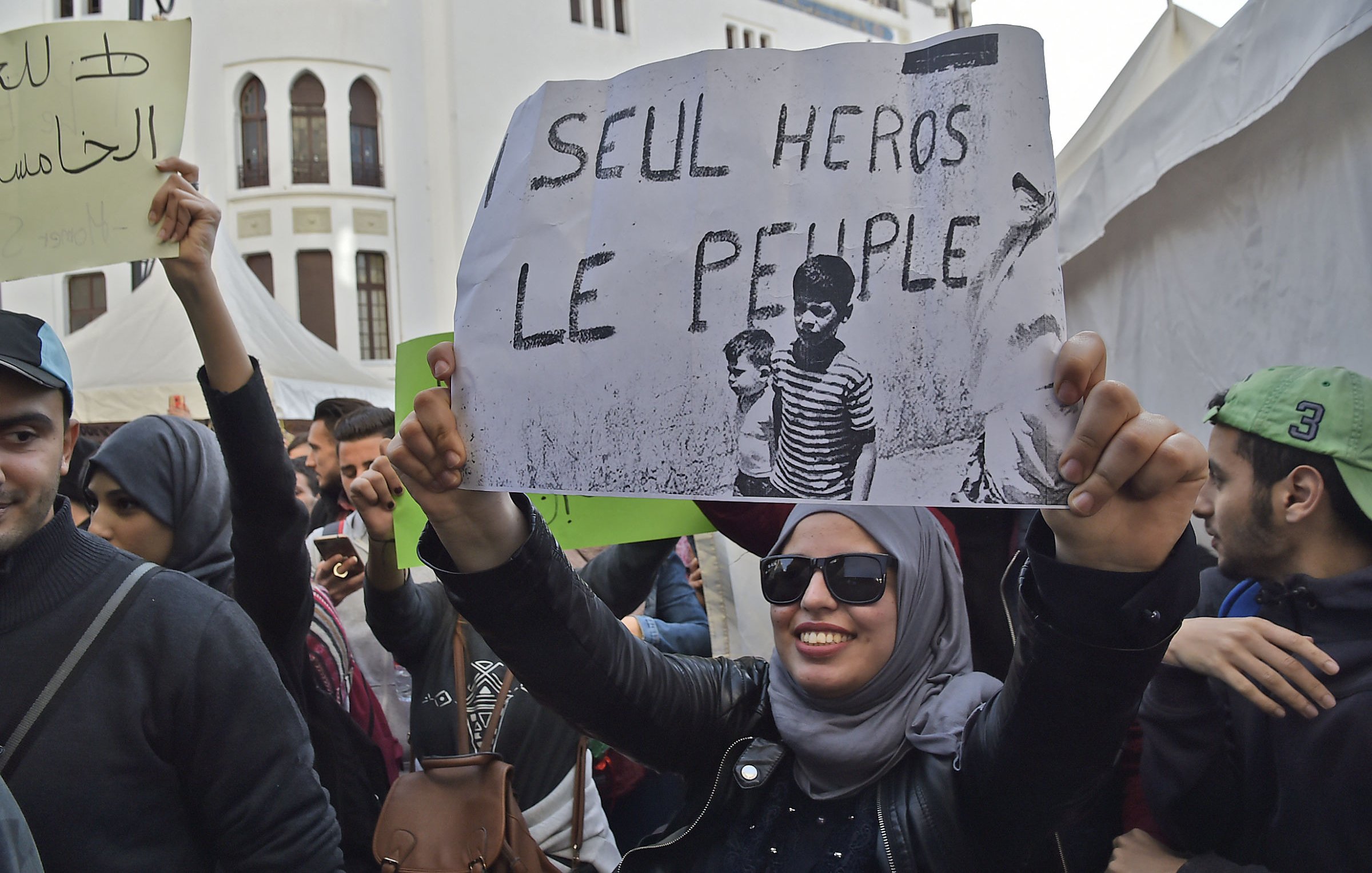 L'image montre une manifestation avec des personnes rassemblées. Au premier plan, une femme portant un hijab et des lunettes de soleil tient une pancarte qui affiche le slogan « SEUL HÉROS LE PEUPLE ? ». L'ambiance semble être engagée, avec d'autres manifestants en arrière-plan et des banderoles visibles. Le décor pourrait suggérer une mobilisation sociale ou politique.