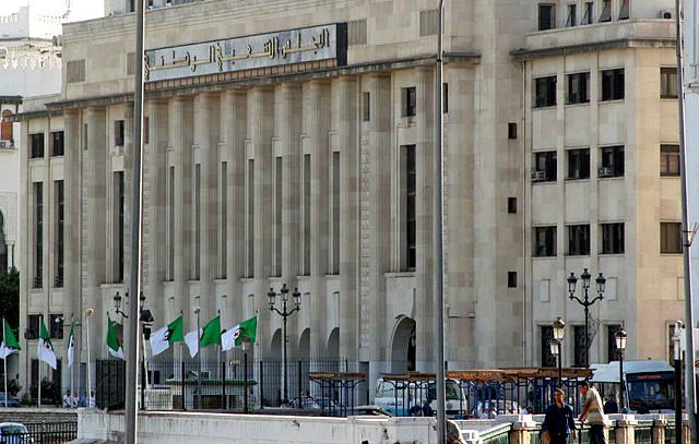 L'image montre un bâtiment majestueux avec une architecture moderne. La façade est claire, avec de grandes fenêtres et des lignes géométriques. On peut également voir des drapeaux algériens flottant devant le bâtiment. Il y a des lampadaires et une ambiance urbaine. Ce contraste entre le style architectural et le décor environnant suggère un lieu important, probablement lié aux institutions gouvernementales.