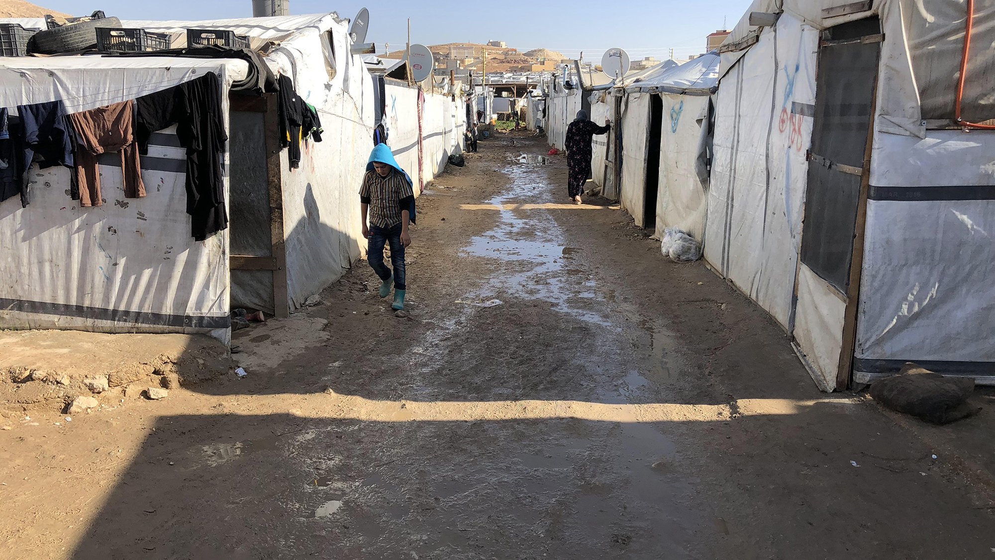 تُظهر الصورة منظرًا لمخيم يتكون من خيام بيضاء متراصة على جانبي ممر ضيق. يبدو أن الأجواء قد كانت ممطرة، حيث تتجمع برك صغيرة من الماء على الأرض. يوجد أشخاص يسيرون في الممر، وبعض الملابس معلقة على الحبال خارج الخيام. يسيطر الشعور بالبساطة والصرامة على البيئة المحيطة، مما يعكس ظروف الحياة في المخيم.