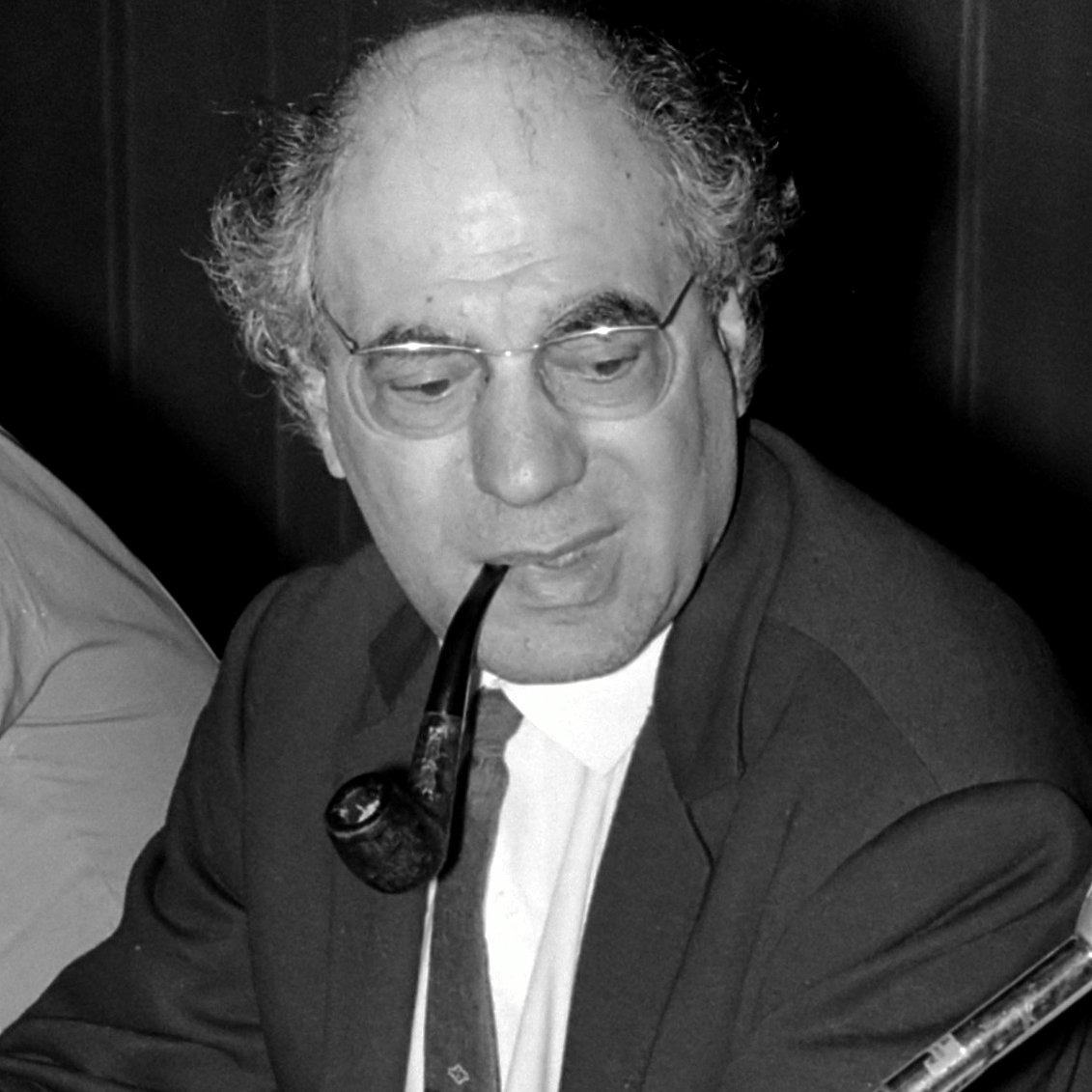 The image features a man seated at a table, wearing a suit and tie. He has a full head of curly hair and is holding a pipe in his mouth, which gives him a thoughtful or contemplative appearance. The setting seems to be a formal or professional environment, likely during a meeting or discussion. The lighting is subdued, which adds to the serious tone of the scene.