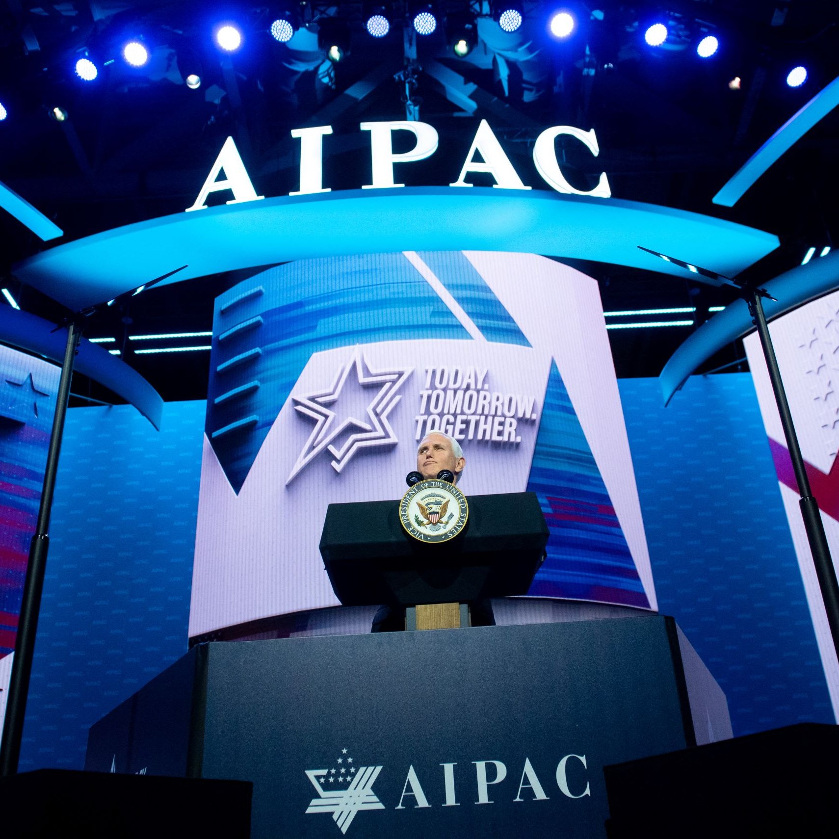 La imagen muestra un escenario iluminado con luces brillantes en un evento, con un gran fondo donde se puede ver el logo de AIPAC (Comité de Relaciones Públicas América-Israel). Hay un podio en el centro, probablemente destinado a un orador, con un sello oficial, indicando que se trata de un evento importante. La decoración incluye elementos gráficos como estrellas y símbolos que refuerzan un mensaje de unidad y futuro, destacado en el fondo.