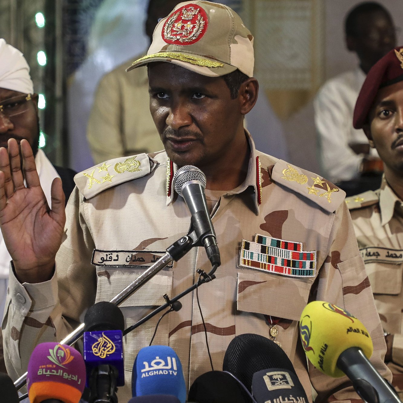 L'image montre un homme en uniforme militaire, portant une casquette décorée, qui s'exprime devant un micro lors d'une conférence de presse. Il semble être en train de faire une déclaration importante, avec une main levée. En arrière-plan, deux autres hommes sont également présents, l'un portant un chapeau traditionnel et l'autre en uniforme militaire. Divers microphones de médias sont placés devant lui. L'ambiance semble sérieuse, suggérant un contexte politique ou militaire.