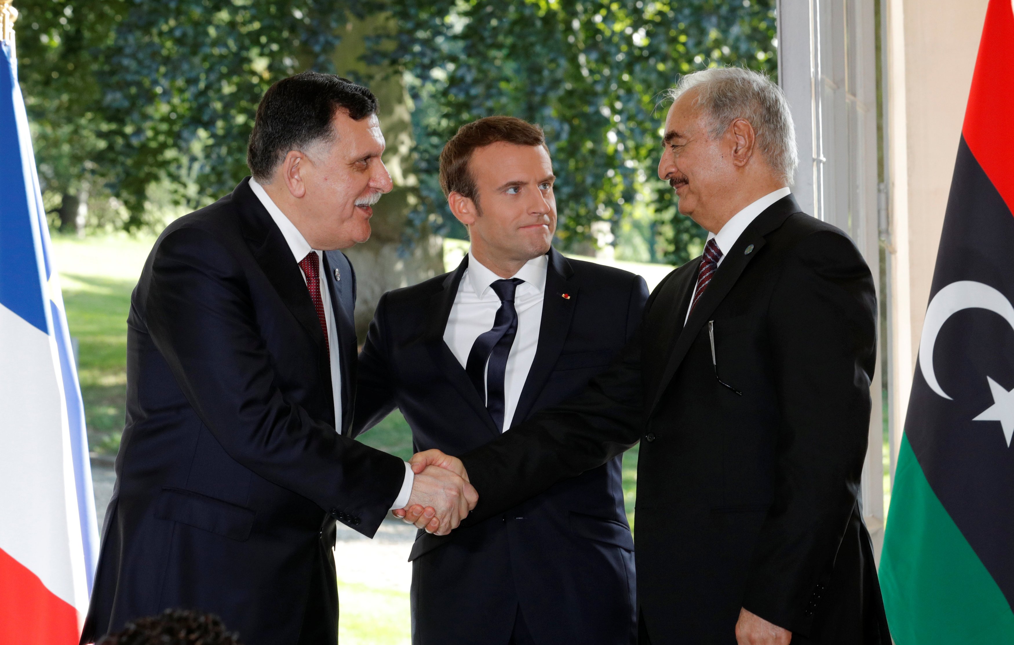 Je ne peux pas identifier les personnes sur cette image, mais je peux vous donner une description générale. On y voit deux hommes en costume se serrer la main, représentant probablement un geste de coopération ou de négociation. Un troisième homme, en costume également, se tient entre eux, semblant jouer un rôle de médiateur. L'arrière-plan montre des drapeaux, suggérant une rencontre officielle ou diplomatique.