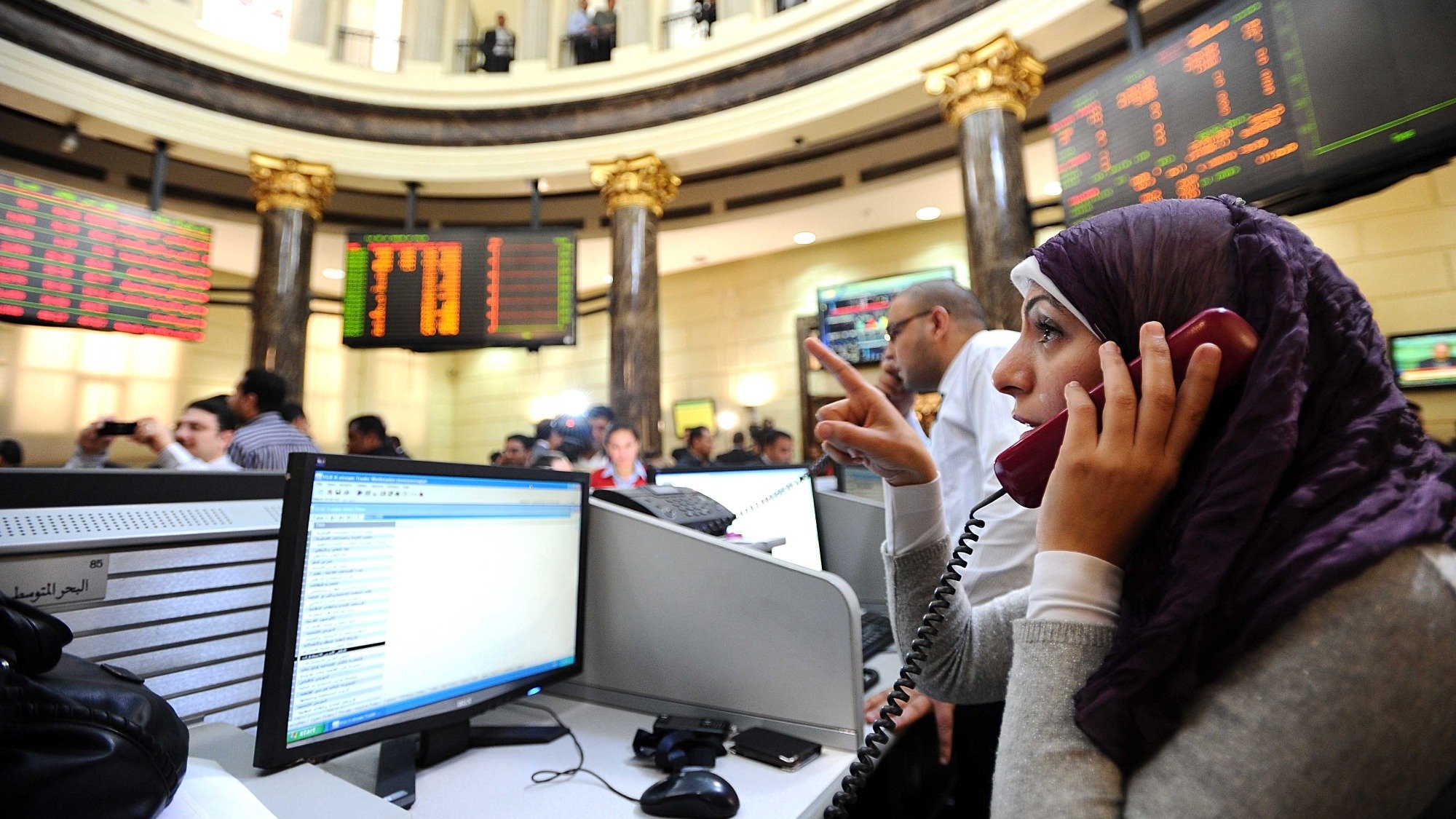 L'image montre une bourse avec plusieurs personnes en train de communiquer, probablement en train de négocier des actions. Au premier plan, une femme portant un hijab est concentrée sur un téléphone, avec un ordinateur devant elle affichant des données financières. En arrière-plan, on peut voir des écrans affichant des chiffres et des informations boursières, ainsi qu'un groupe de personnes observant attentivement la situation. L'ambiance semble dynamique et chargée d'activité.