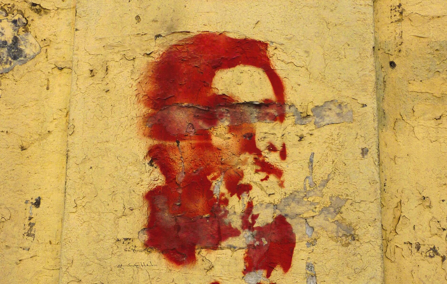 L'immagine mostra un murale su una parete di colore giallo. Il murale rappresenta un volto stilizzato, probabilmente un personaggio noto, dipinto in rosso. Sotto il volto ci sono delle scritte, anch'esse in rosso, che sembrano in arabo. La superficie della parete appare consumata e scrostata, contribuendo a un aspetto storico e di protesta.