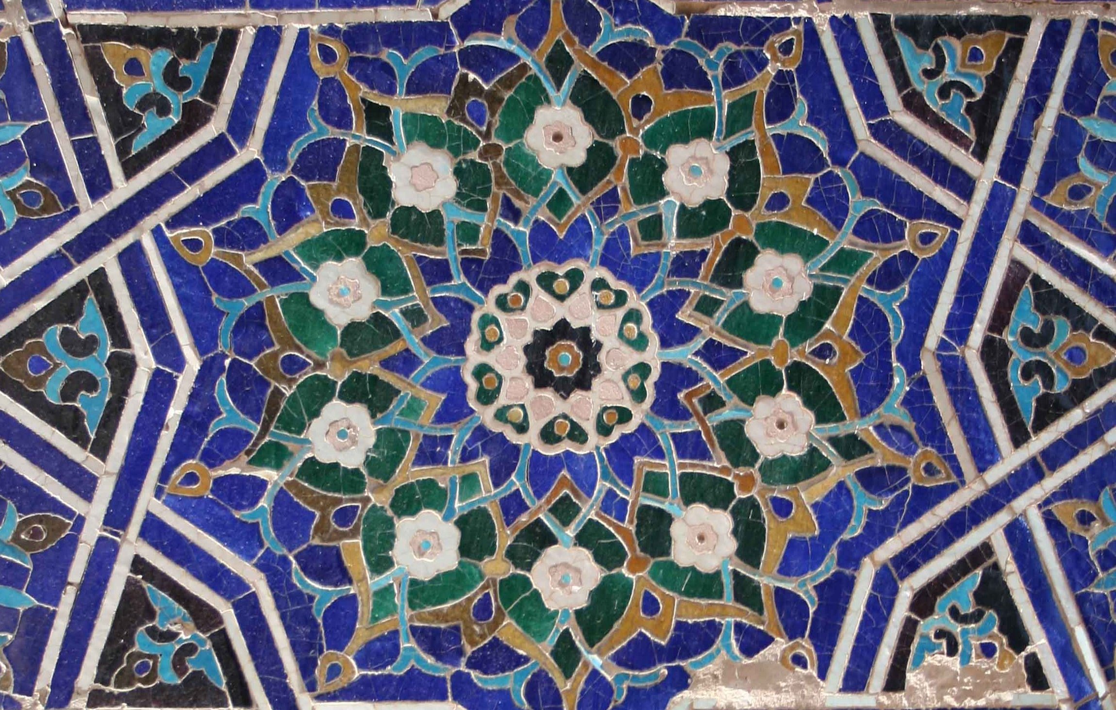 The image features an intricate geometric design characterized by vibrant colors, including deep blues, greens, browns, and whites. At the center, there is a floral motif surrounded by symmetrical patterns that radiate outward. The design showcases a combination of star shapes and circular forms, typical of traditional tile work found in Islamic art. The use of intricate detailing and color contrasts adds depth and visual appeal to the overall composition.