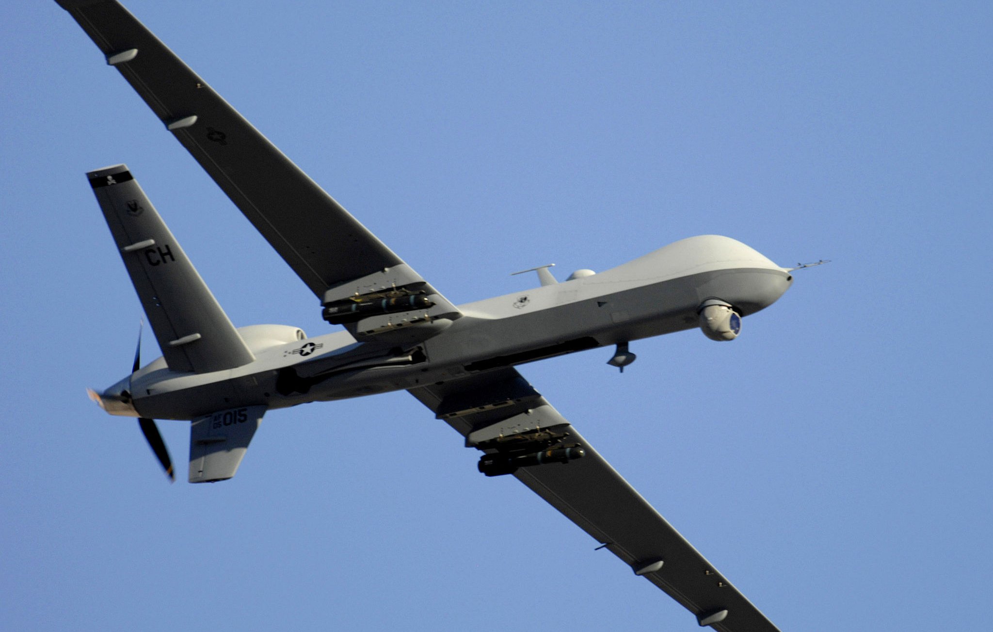 L'image montre un drone de type militaire, en vol. Il a une conception aérodynamique avec des ailes étendues et une queue distinctive. La surface du drone est peinte en gris, ce qui le rend moins visible en altitude. On peut également observer des équipements sous ses ailes, probablement destinés à la surveillance ou à des missions de reconnaissance. Le ciel en arrière-plan est dégagé, mettant en valeur le drone en mouvement.