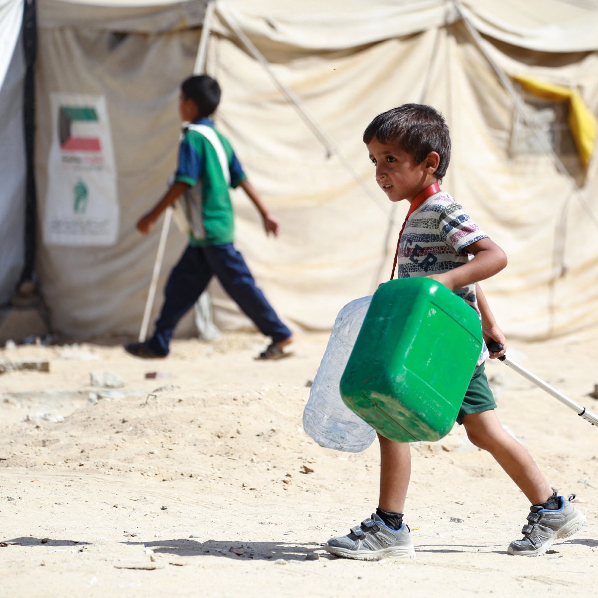 در تصویر، پسربچه‌ای دیده می‌شود که در حال حمل یک کوله‌پشتی به رنگ صورتی و یک ظرف سبز رنگ است. او در یک محیط بیابانی و خشک قرار دارد که چادرهای موقتی در پس‌زمینه به چشم می‌خورد. پسربچه لباسی ساده و راحت پوشیده و به نظر می‌رسد که در حال حرکت به سمت مقصدی است. در کنار او، کودک دیگری نیز در حال گذر است. فضای تصویر حالتی از زندگی در شرایط سخت را به نمایش می‌گذارد.