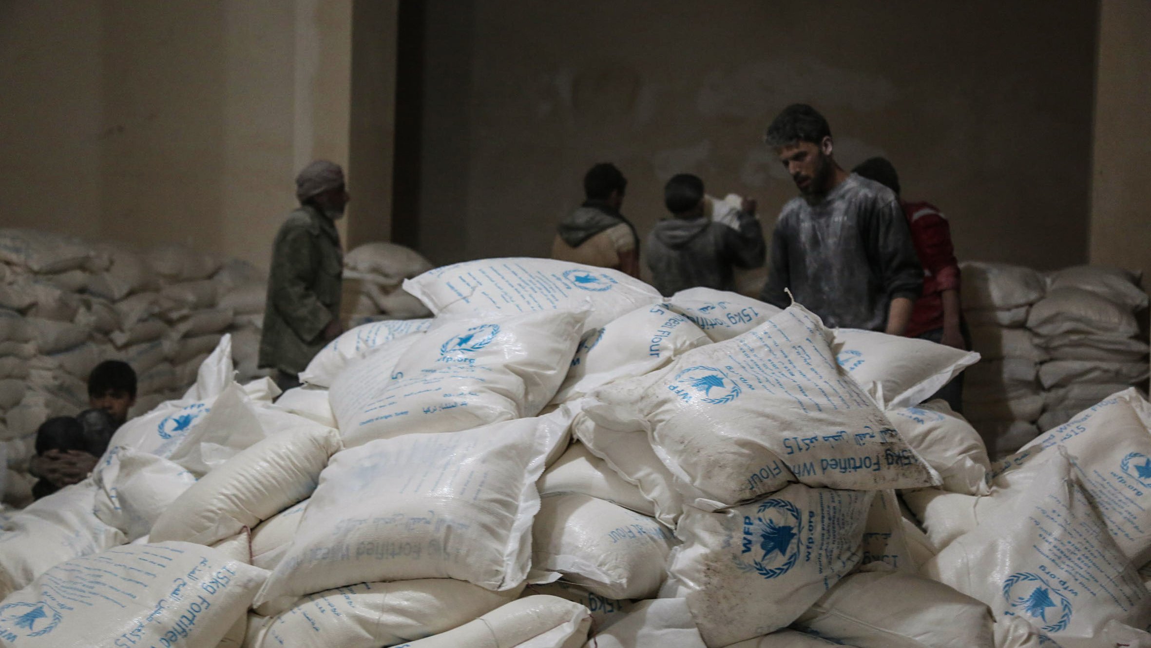 تظهر في الصورة مجموعة من الأشخاص داخل مكان يبدو أنه مخزن للمواد الإغاثية. في المقدمة، هناك كومة كبيرة من الأكياس البيضاء التي تحمل علامات معينة، ويبدو أن الأكياس تحتوي على مواد غذائية أو إغاثية. في الخلفية، يمكن رؤية بعض الأشخاص الذين يتحدثون أو يعملون. الإضاءة في المكان خافتة، مما يعطي شعورًا بالجدية والاهتمام بالعمل الذي يقومون به.