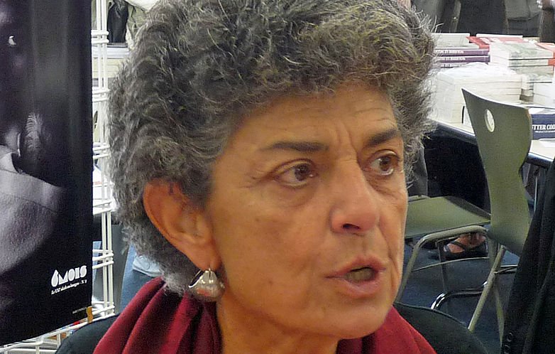 Je ne sais pas qui est la personne sur l'image. Cependant, je peux décrire l'apparence générale : il s'agit d'une femme avec des cheveux gris et bouclés, portant un gilet noir et une écharpe rouge. Elle semble engagée dans une conversation, exprimant des émotions. En arrière-plan, on distingue des personnes et des livres.