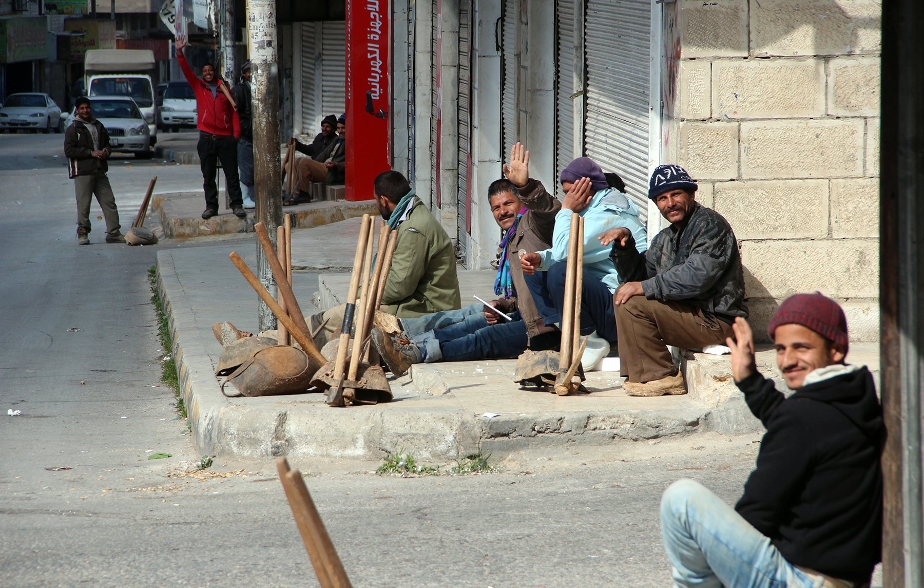 الصورة تظهر مجموعة من الرجال يجلسون على الرصيف في منطقة حضرية. بعضهم يلوح بيده وكأنهم يتحدثون أو يتبادلون الحديث. يبدو أن لديهم أدوات عمل قربهم، مما يوحي بأنهم ربما كانوا ينتظرون فرصة للعمل. الشارع يبدو هادئًا ومظهراً لأبنية متوسطة الارتفاع. الأجواء تعكس نشاطًا ونمط حياة يومي في المدينة.