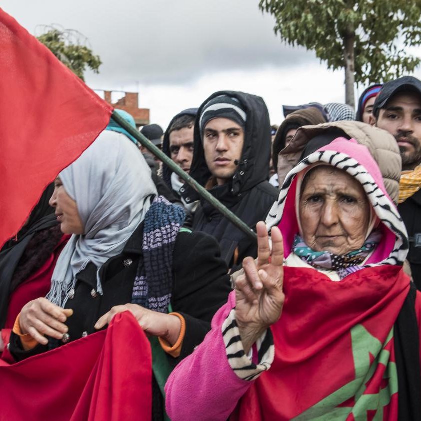 تظهر الصورة مجموعة من الأشخاص يتجمعون في مظاهرة أو احتفال. في المقدمة، هناك امرأة مسنّة تحمل علم المغرب، وتُظهر إشارة النصر بإصبعها. يرتدي بعض المشاركين في الخلف ملابس متنوعة، ومن بينهم شباب ونساء. الأعلام الحمراء التي تحمل رمز المغرب تُظهر الفخر والهوية الوطنية، بينما تبدو الأجواء مليئة بالحماس والترقب. السماء تبدو غائمة، مما يشير إلى تغيرات محتملة في الطقس.