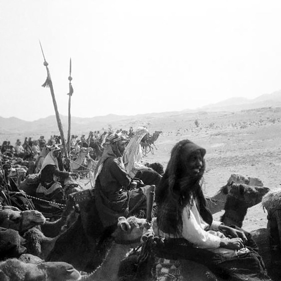 تصویری سیاه و سفید از یک کاروان در بیابان است. گروهی از افرادی که بر روی شترها سوار هستند، در حال حرکت به سمت دوربین هستند. در میان آنها زنانی با پوشش‌های سنتی و مردانی با لباس‌های مختلف دیده می‌شوند. برخی از افراد نیز پرچم‌هایی در دست دارند. پس‌زمینه تصویر شامل تپه‌های شنی و کوه‌ها است که حس فضای بیابانی را القا می‌کند. جو تصویر قدیمی و تاریخی به نظر می‌رسد.