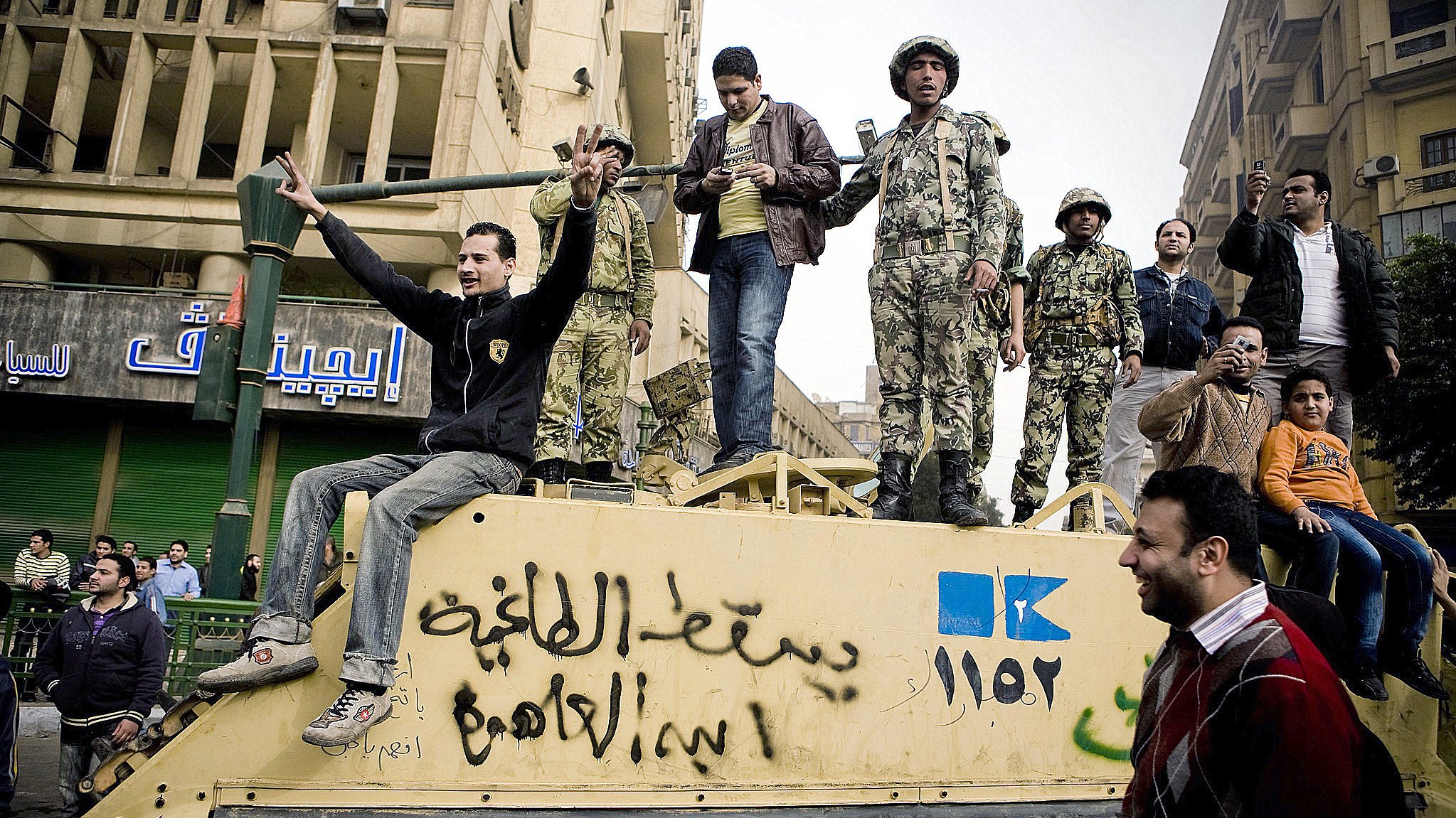 تظهر الصورة مجموعة من الأشخاص، بعضهم يرتدي زيًا عسكريًا، بينما الآخرون يرتدون ملابس عادية. يبدو أن هؤلاء الأشخاص متواجدون فوق مركبة عسكرية أو دبابه، حيث يعبر البعض عن مشاعرهم بإشارات وأيدي مرفوعة. يحاط بهم بعض المدنيين الذين يدفعون بحماس. الخلفية تحتوي على مباني وشوارع تشير إلى حضرية البيئة المحيطة. يمكن أن تعكس الصورة أجواء من التوتر أو الاحتجاجات.