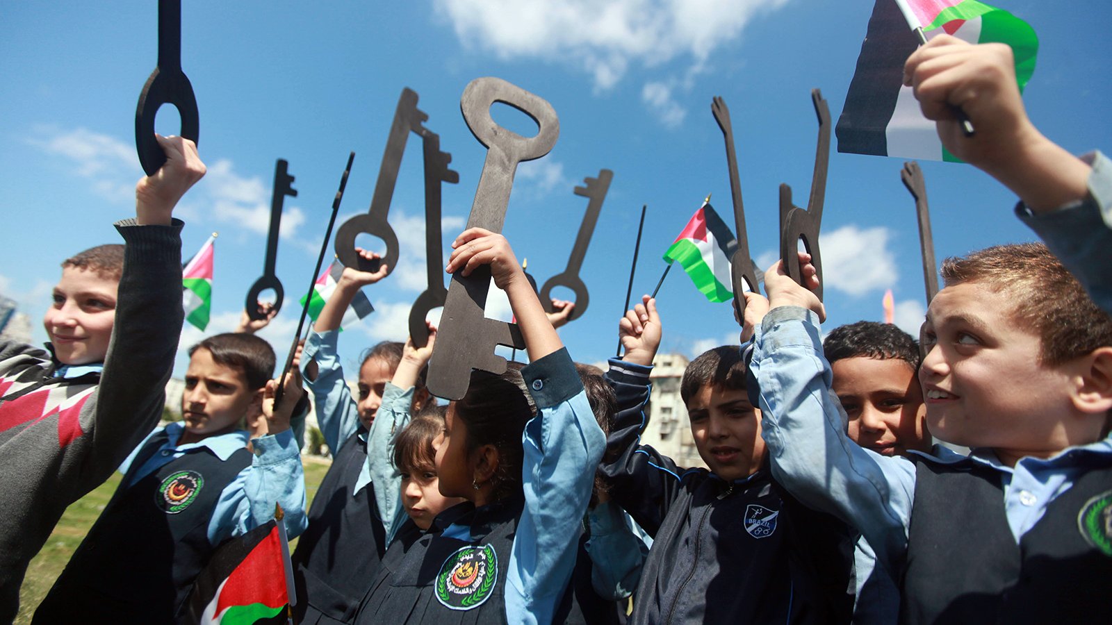 L'image montre un groupe d'enfants, qui semblent être en train de célébrer ou de manifester. Ils tiennent des grandes clés en carton et des drapeaux palestiniens. Les enfants, vêtus de uniformes scolaires, sourient et lèvent les clés vers le ciel, symbolisant un message de revendication ou d'espoir. Le ciel est bleu avec quelques nuages, ce qui ajoute à l'ambiance de la scène.